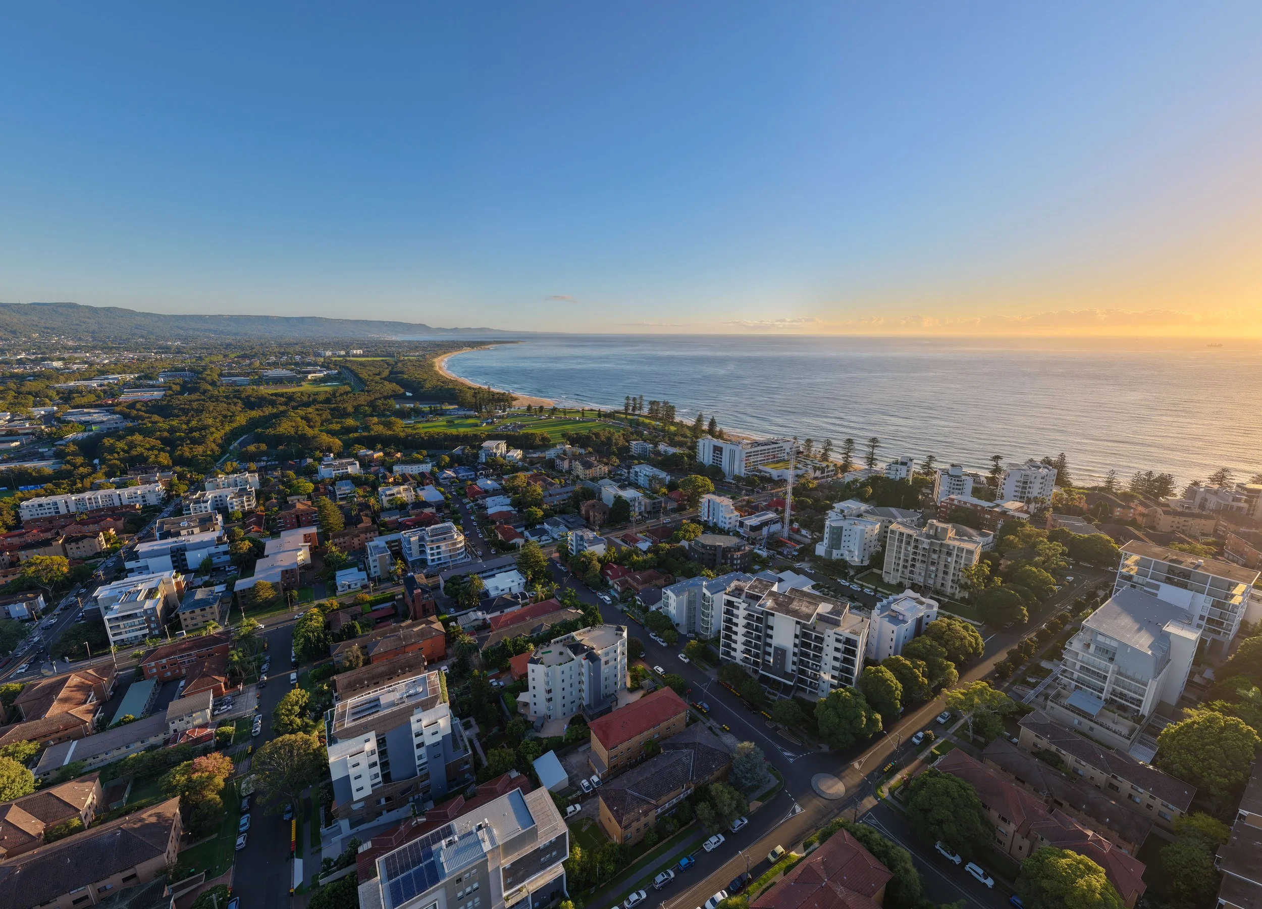 An aerial view of a Wollongong during sunrise, featuring residential buildings, roads, green trees, and a beach along the ocean.