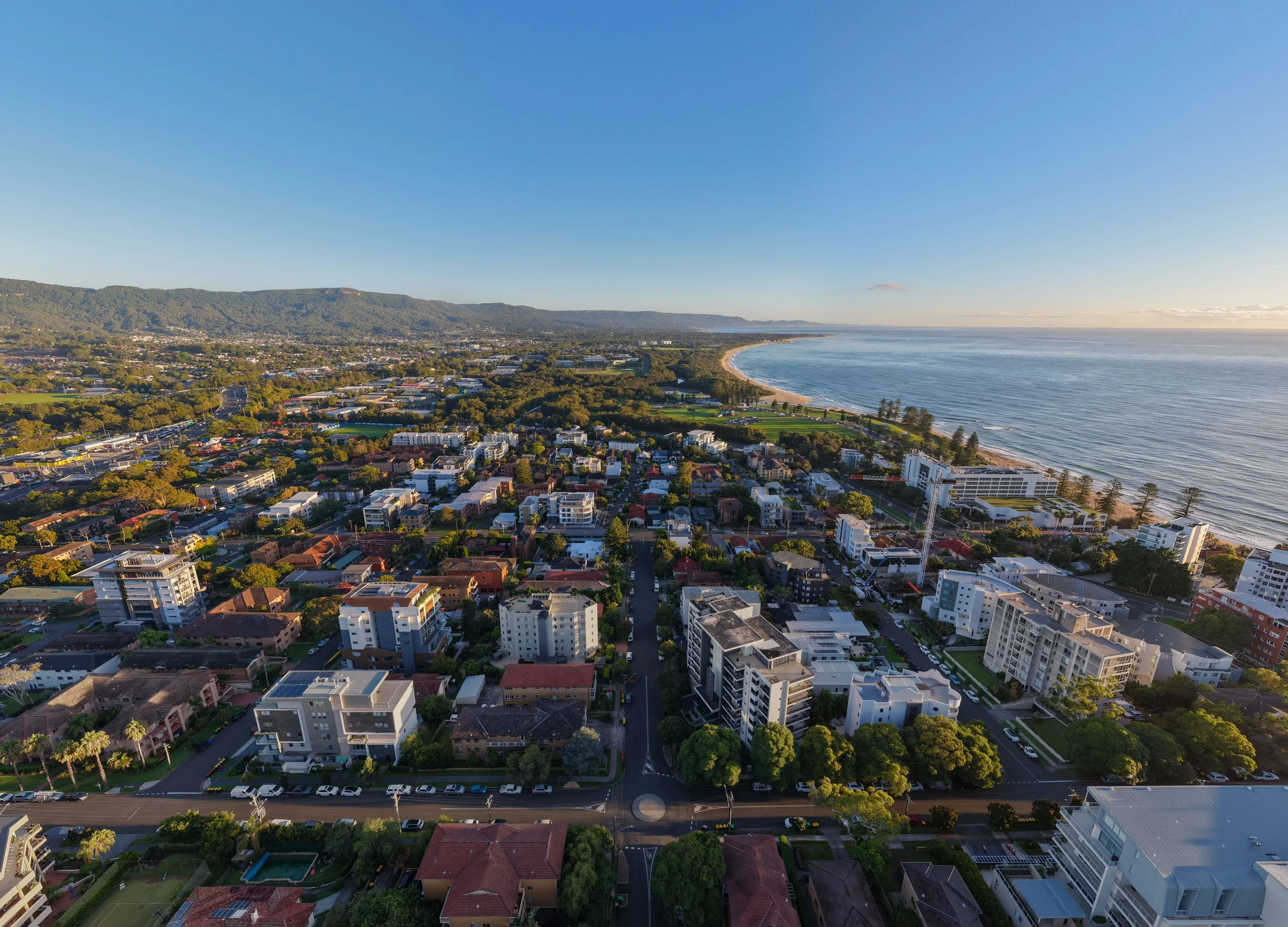 Aerial view of a coastal city with residential and commercial buildings, green trees, a shoreline with a beach, and the ocean extending to the horizon during sunset.