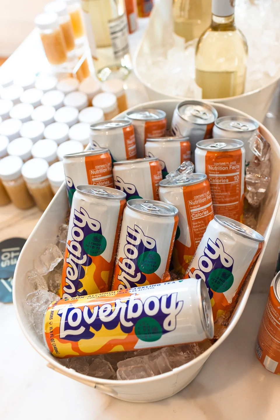 A white bowl filled with ice holding cans of Lemon LoveTown soda. In the background, there are bottles and containers, possibly of beverages or syrups, on a counter.