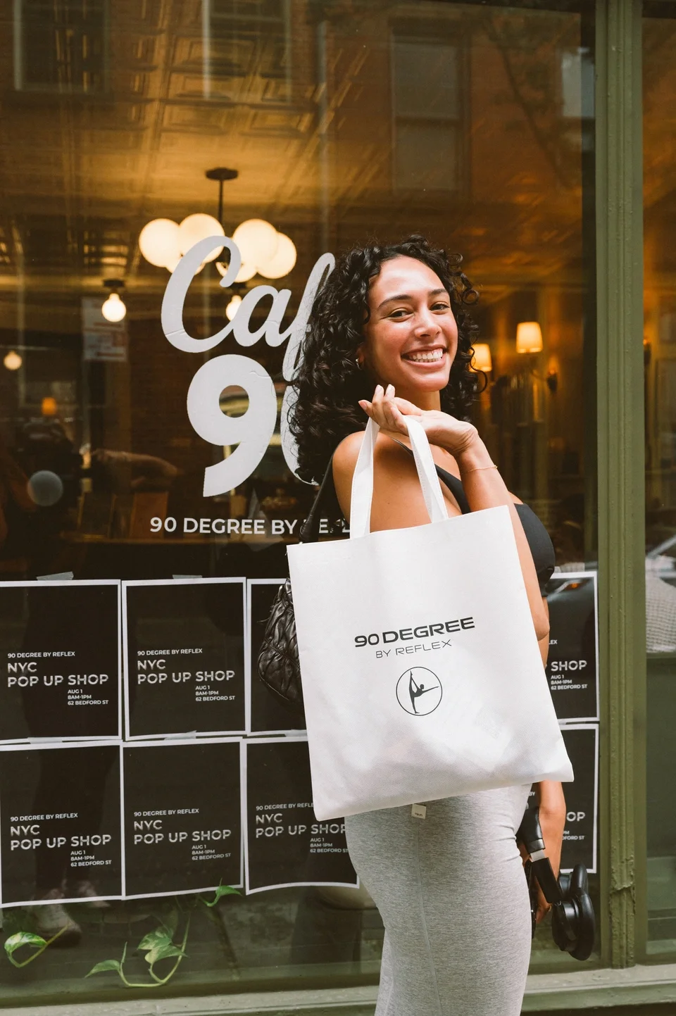 A smiling woman holds a white shopping tote with '90 Degree by Reflex' written on it outside a shop window. The window displays the store name and information about a pop-up shop event. The woman has curly dark hair and is dressed in a black top and gray pants.