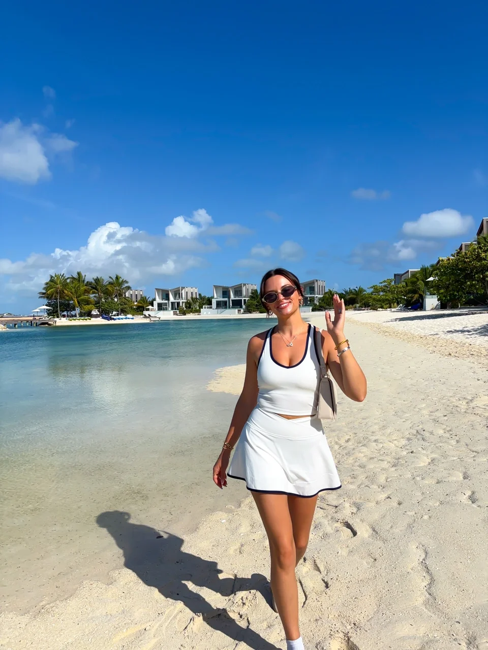 Woman in sunglasses wearing a white tank top and skirt, walking on a sandy beach with clear water, palm trees, and modern buildings in the background under a blue sky with clouds.