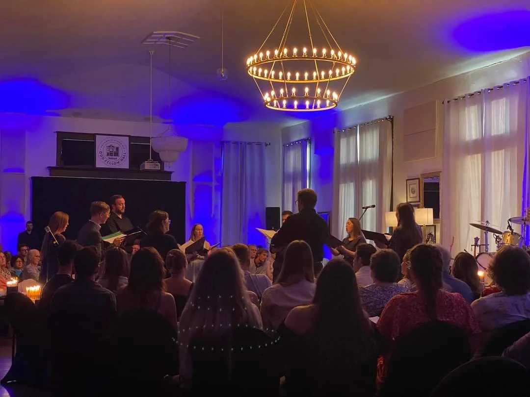 A choir performing indoors with a conductor leading. Audience seated, some candles on tables, elegant chandelier, and blue ambient lighting.