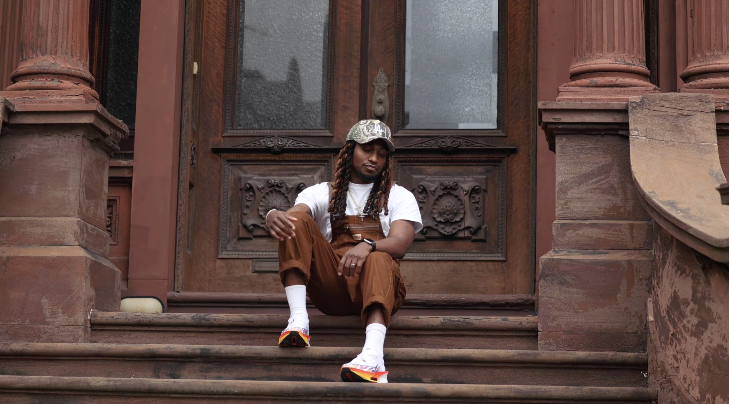 The DreamR with long, braided hair wearing a camo baseball cap, white T-shirt, brown overalls, white socks, and colorful sneakers, sitting on steps in front of a large, ornate wooden door with decorative carvings.