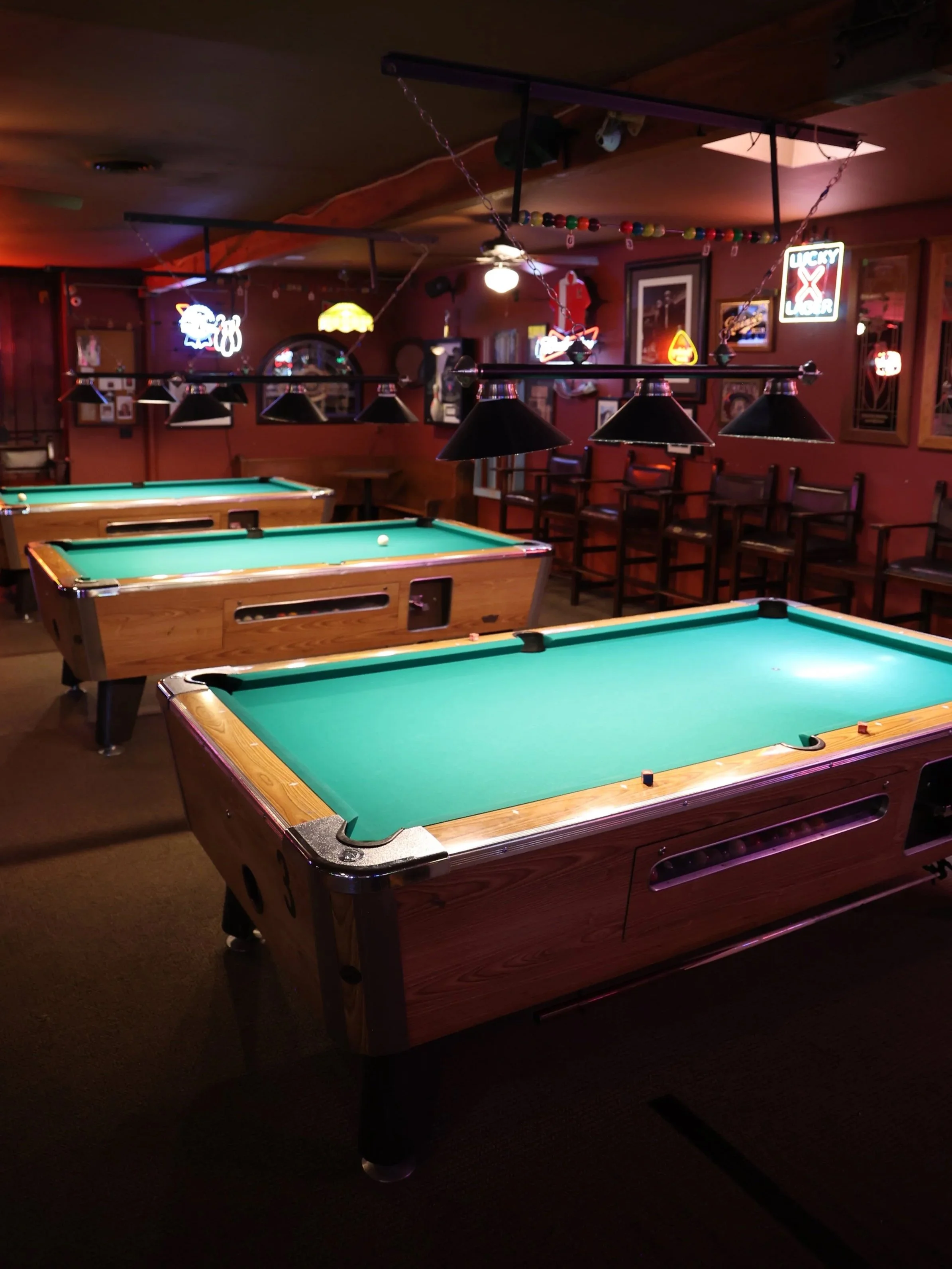 Dimly lit bar with three pool tables, hanging lamps above, neon signs on the walls, and lounge chairs along the back wall.