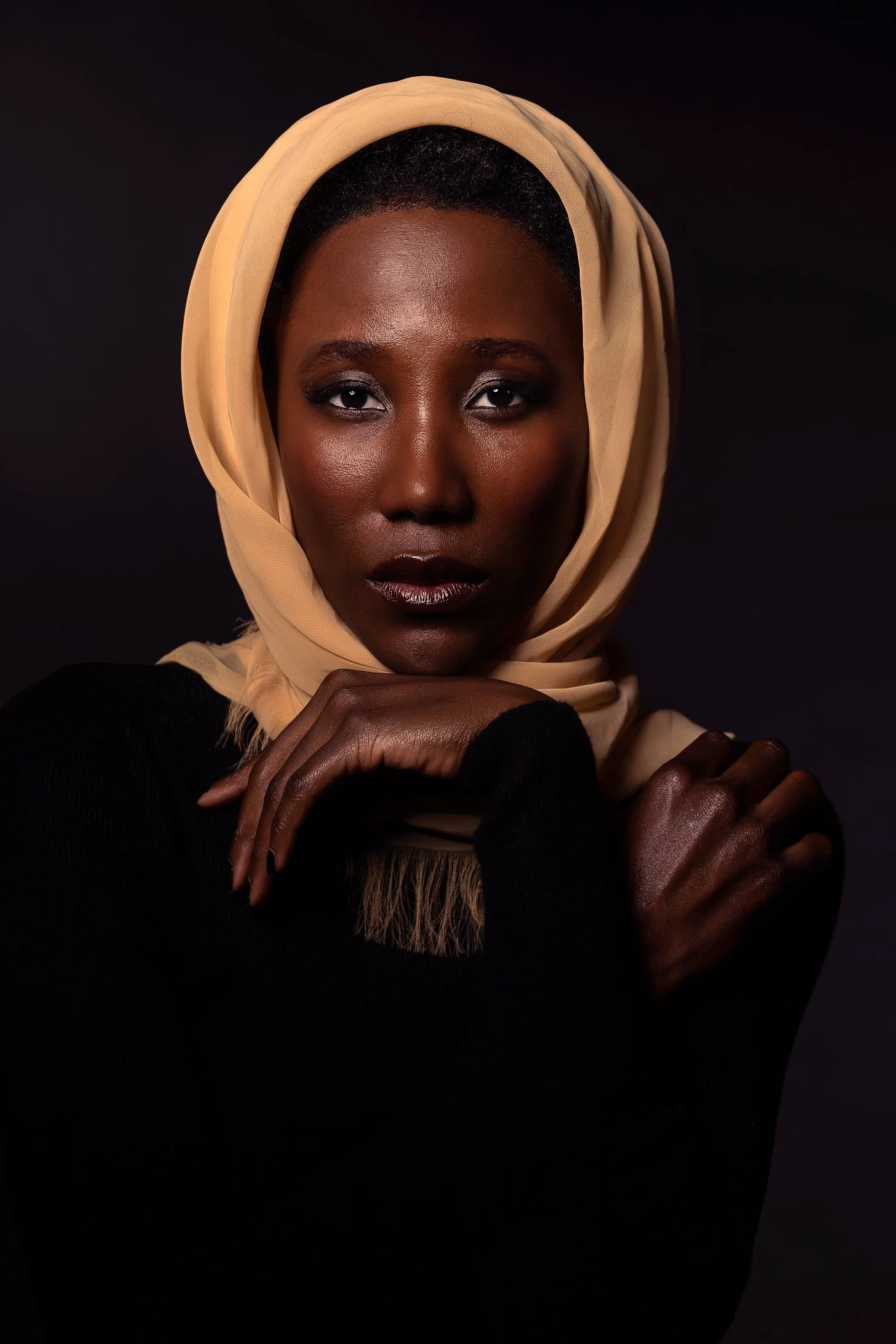 Portrait of an African woman with a beige headscarf, dark lipstick, and dark eye makeup, wearing a black top, posing with crossed arms against a dark background.