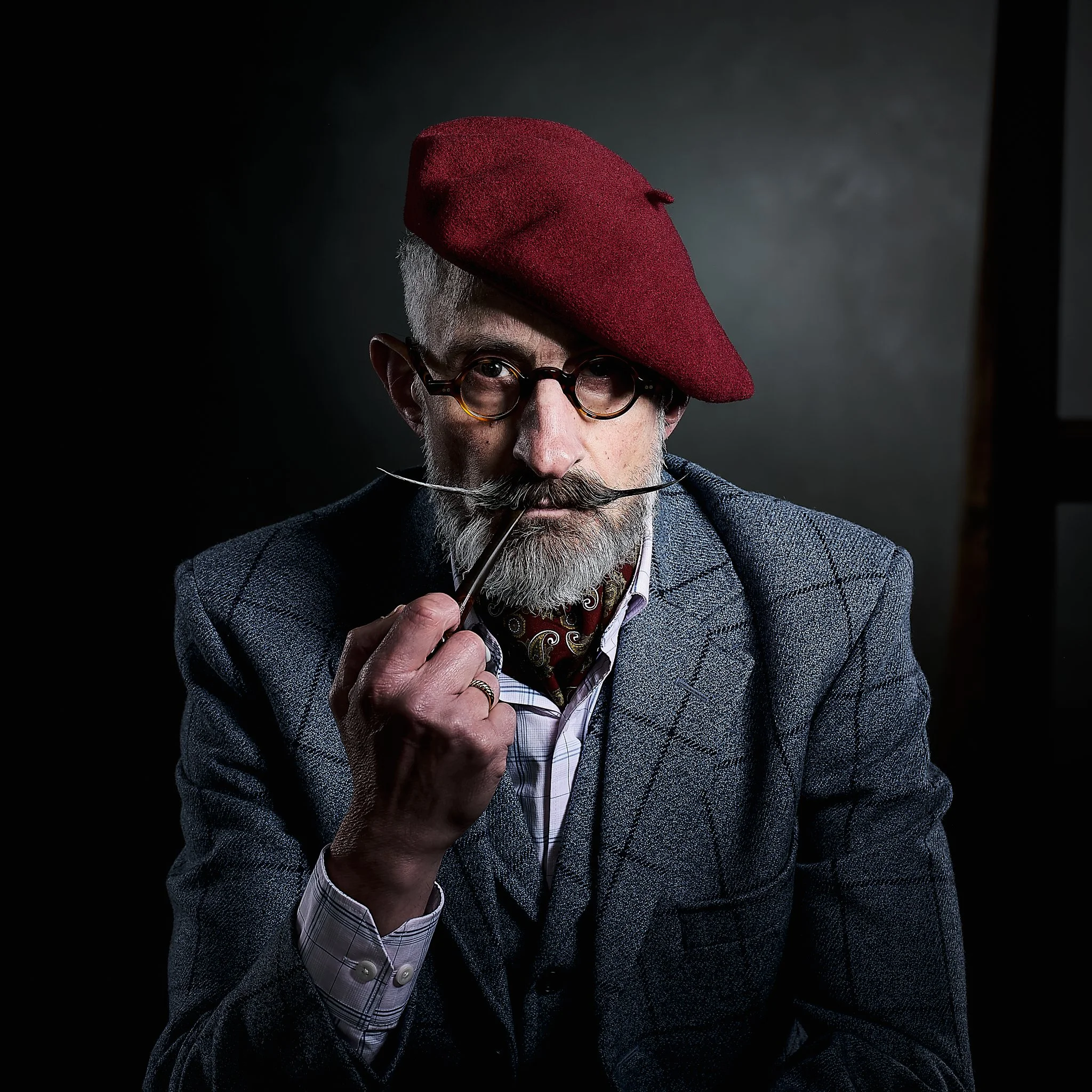 A distinguished elderly man with a grey beard and mustache, wearing glasses and a dark grey checkered suit, holds a pipe near his mouth, and wears a red beret tilted to one side, against a dark background.