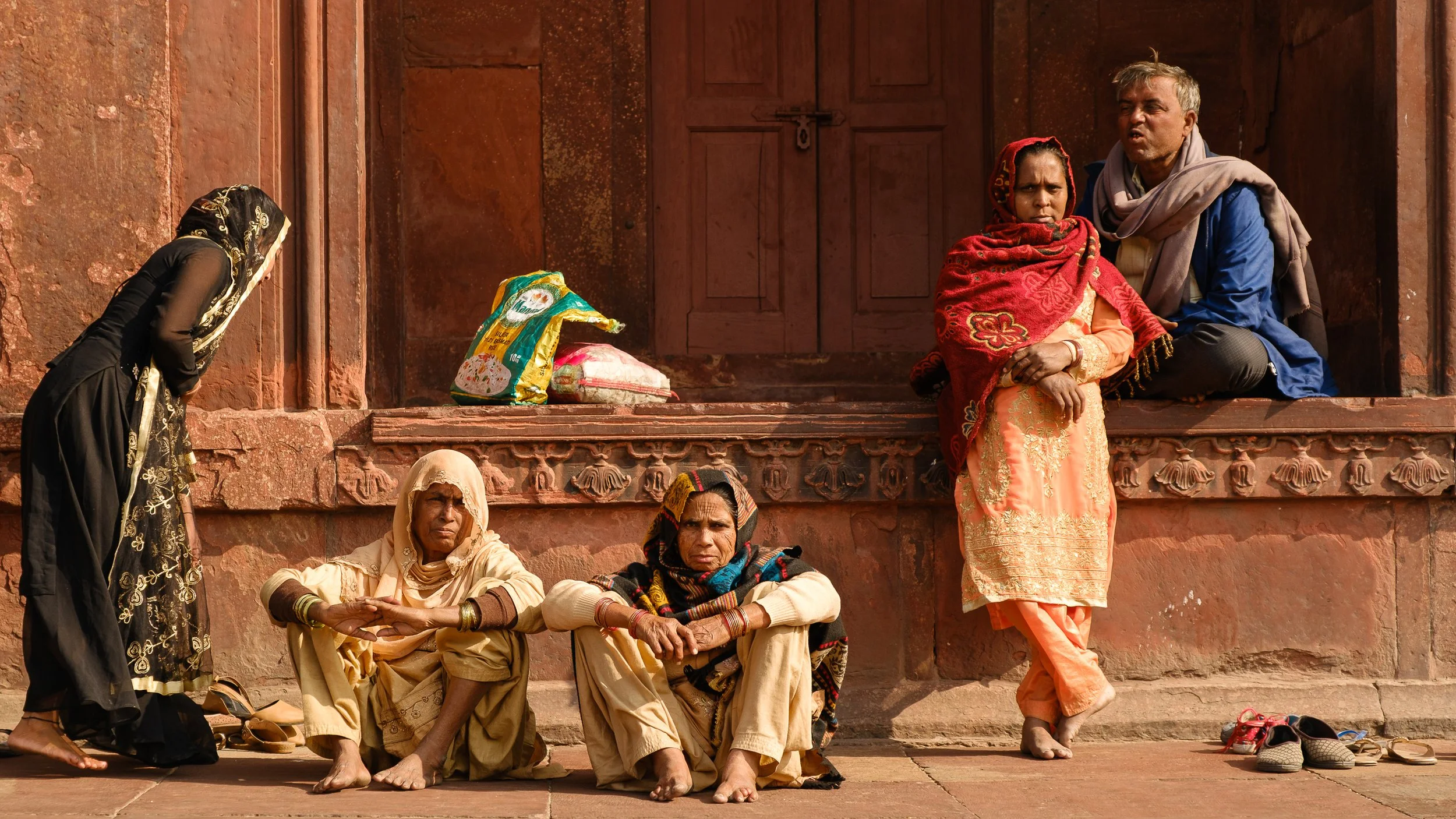 Group of five Indian people outside a building with red sandstone walls; two women sitting on the ground, a woman standing, a man and a woman sitting on a ledge. Shoes are removed and placed on the ground.
