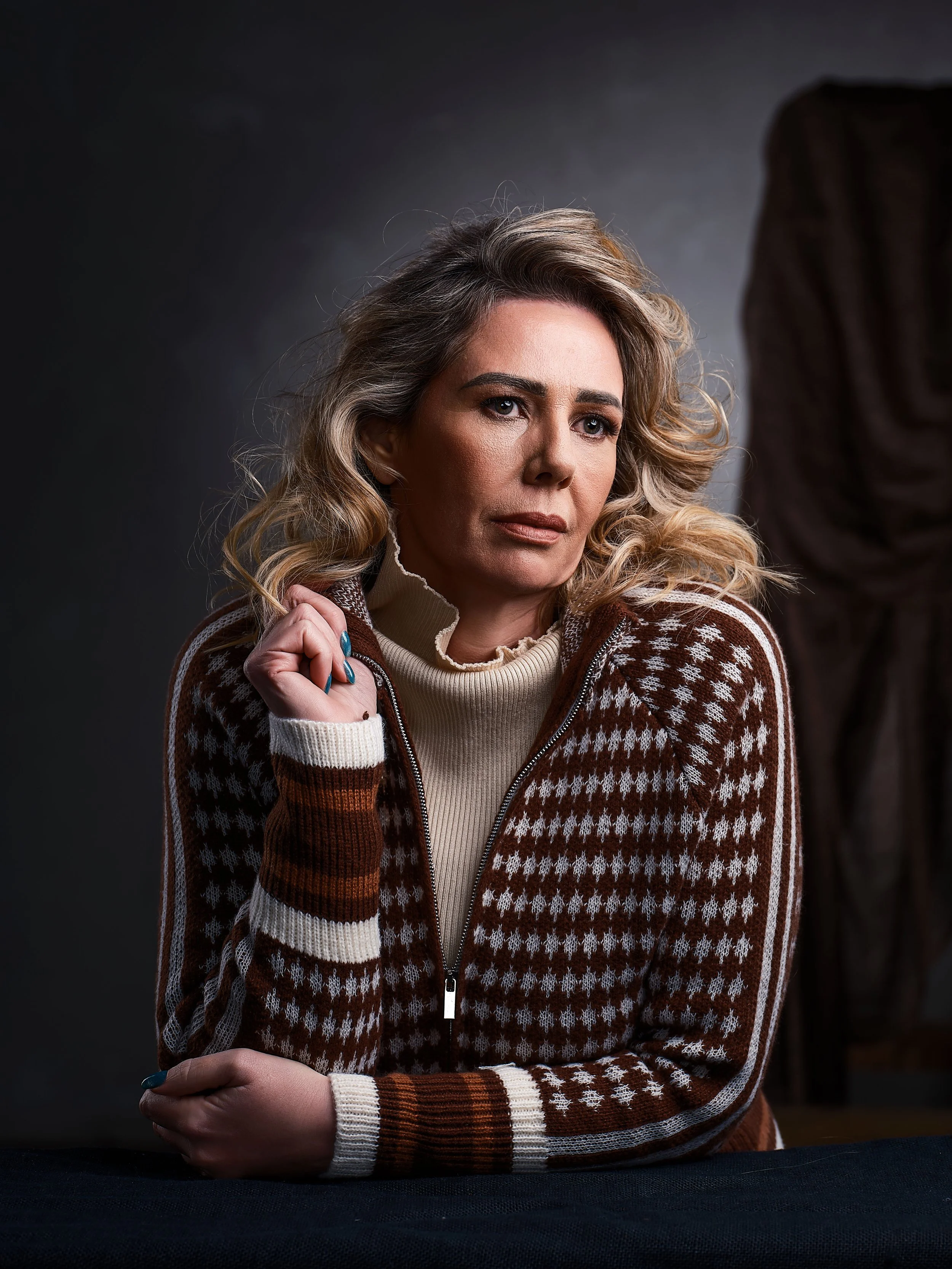 A woman with blonde, shoulder-length wavy hair looking contemplative, wearing a brown and white patterned sweater and a beige turtleneck, seated at a table against a dark background.