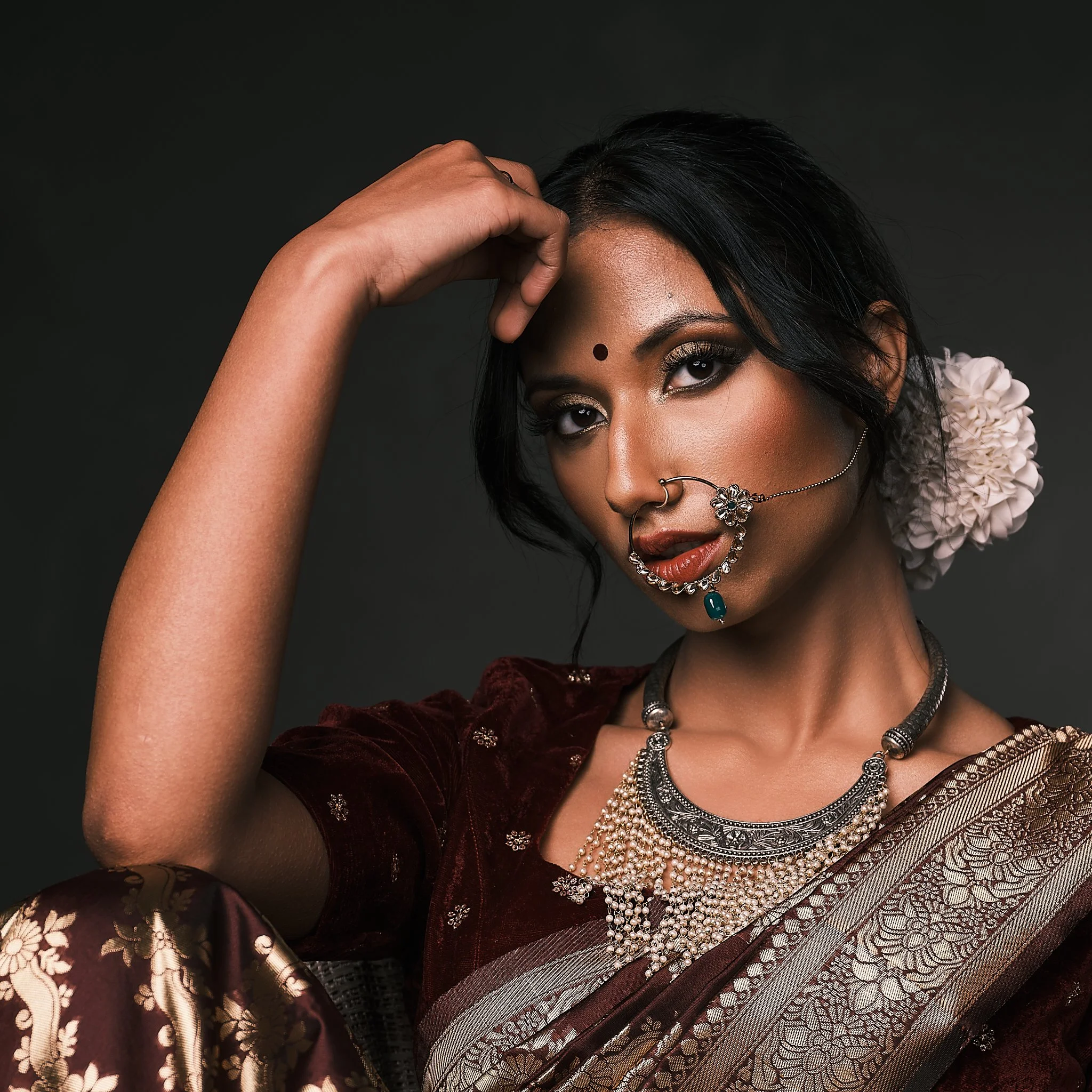 A woman wearing traditional Indian attire and jewelry, with her hand resting on her forehead and a dark background.