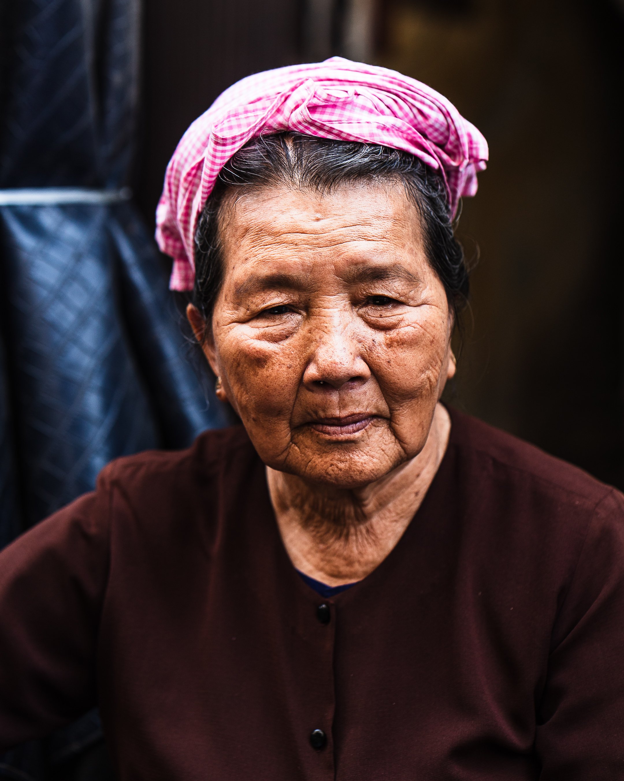 Close-up portrait of an elderly woman with dark hair, wearing a pink and white checkered headscarf and a dark brown top, with a blurred dark background.