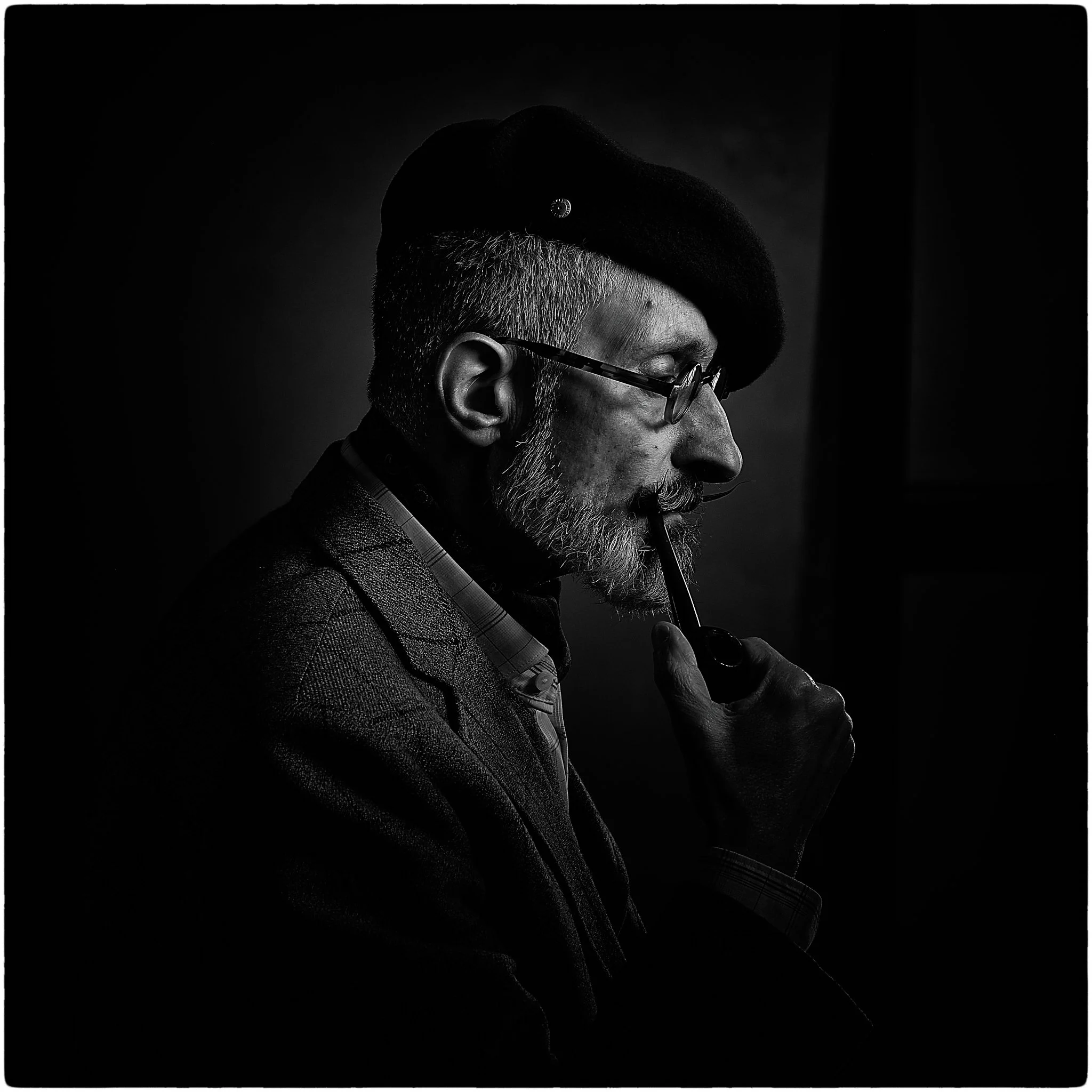 Black and white portrait of an elderly man wearing glasses, a beret, and a plaid jacket, holding a pipe to his lips and looking down thoughtfully.