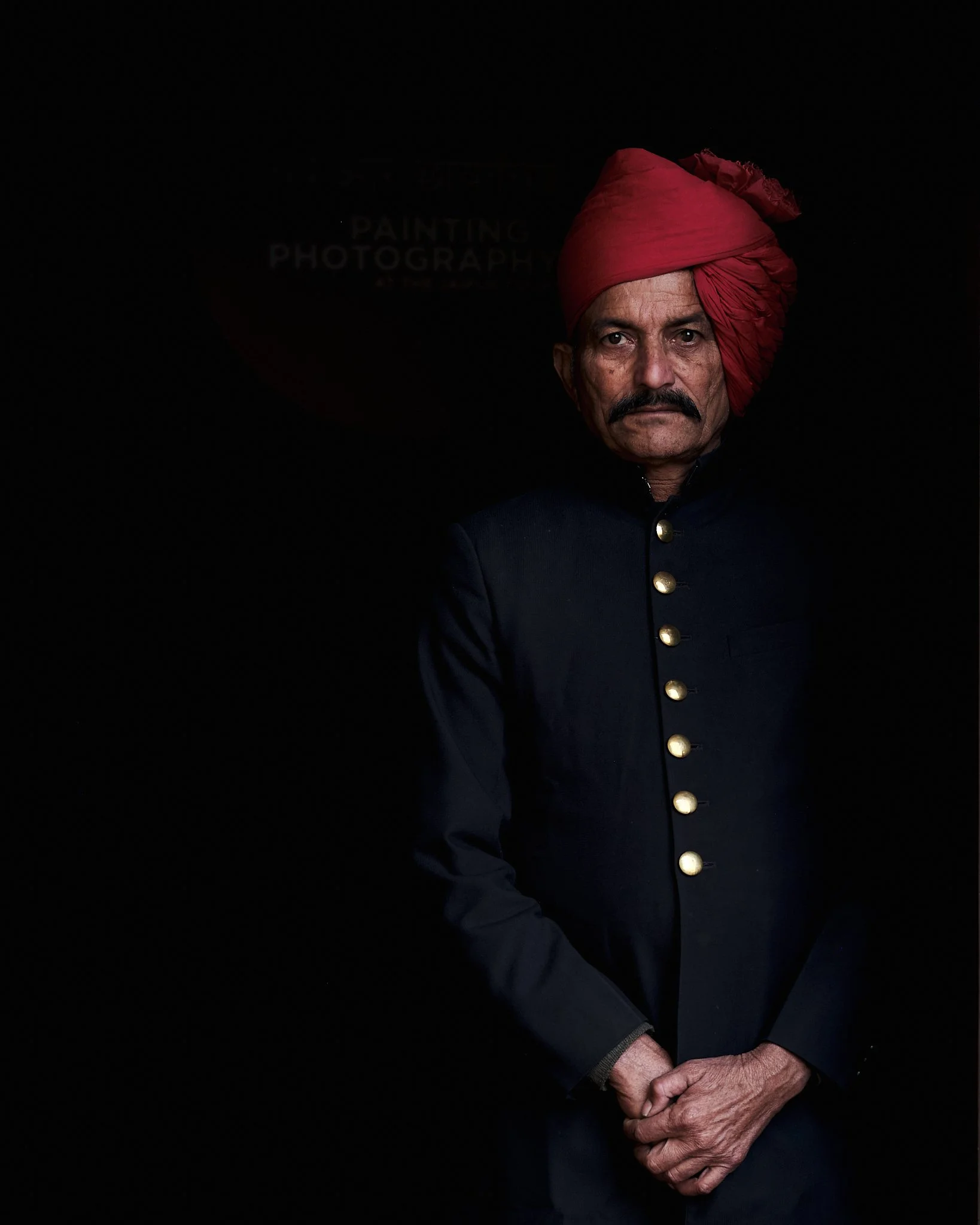 A man wearing a red turban and dark formal traditional Indian attire with brass buttons, standing against a black background.