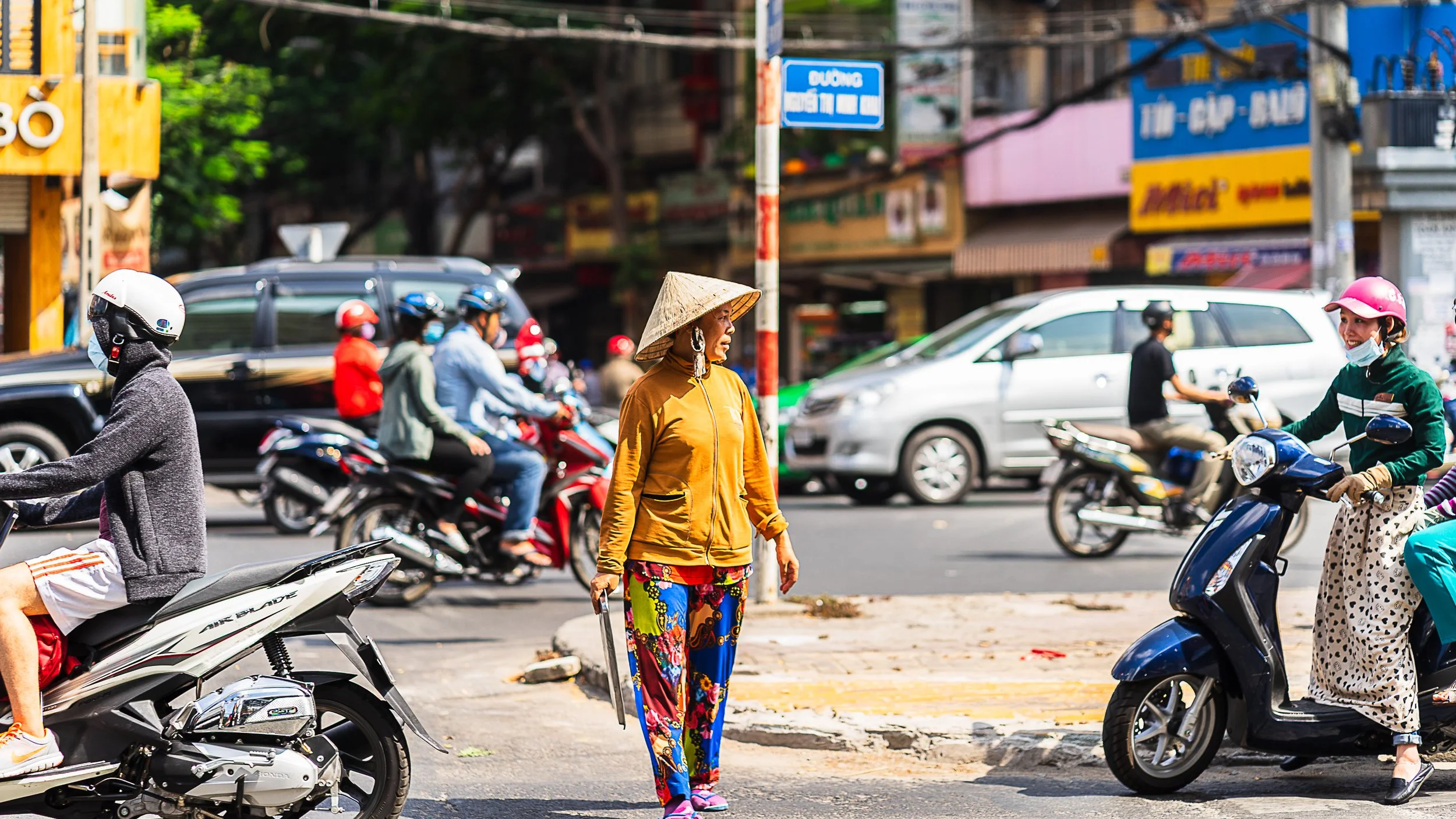 A busy city street scene with multiple motorcyclists and scooters, a woman wearing colorful pants, a yellow jacket, and a traditional conical hat crossing the street, and other vehicles in the background.
