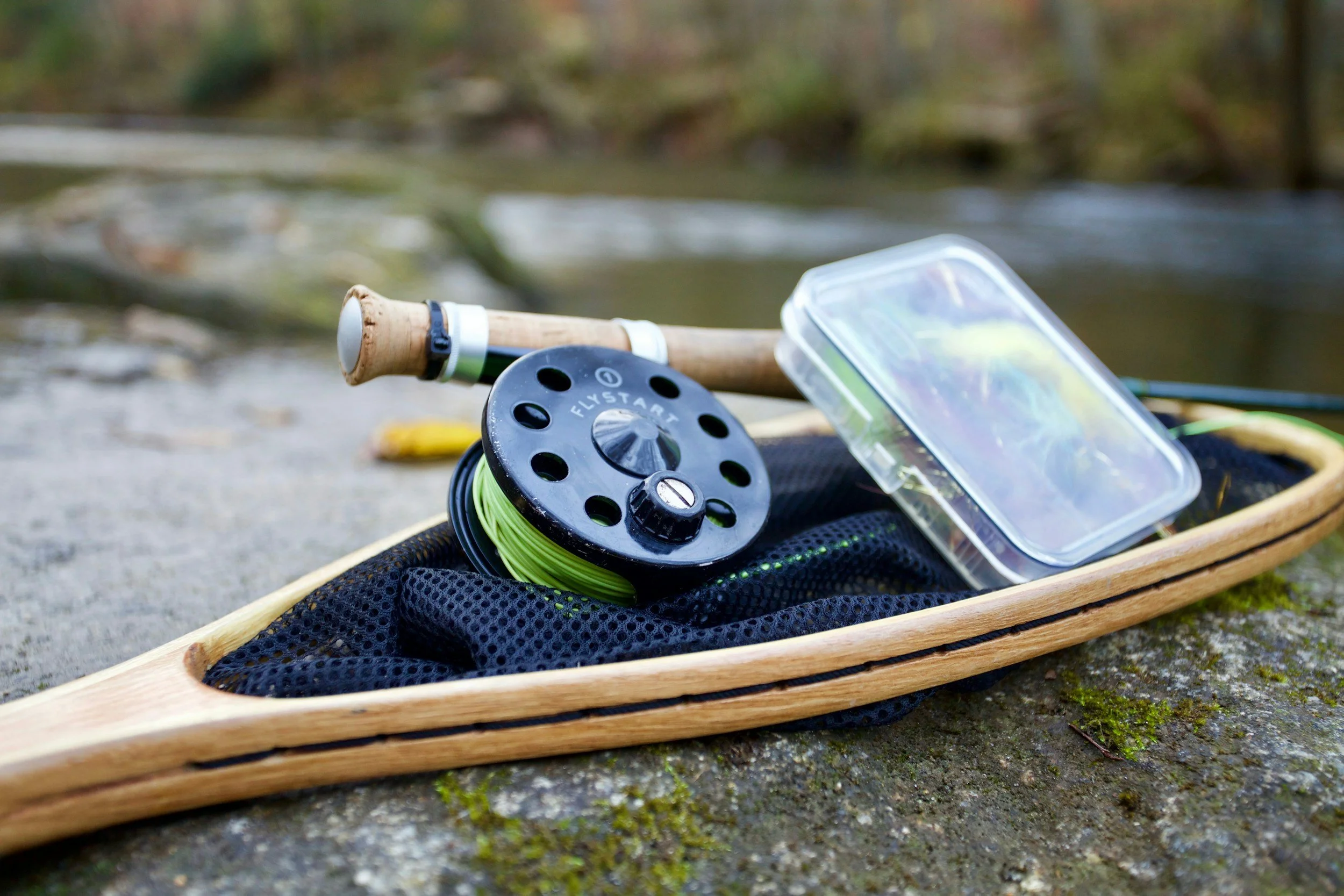 Fishing rod, reel, and tackle box placed on a wooden boat near a riverbank in a wooded area.