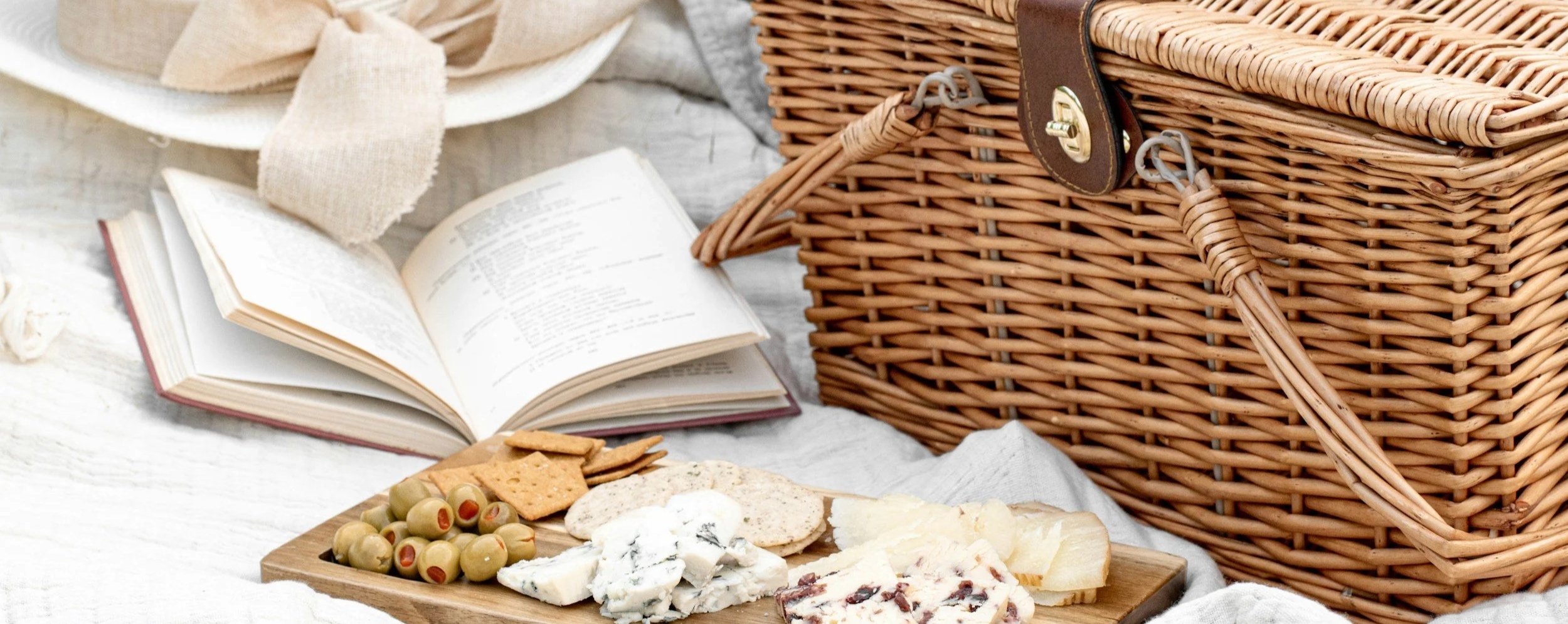 A picnic scene with a wicker picnic basket, an open book, a platter of assorted cheeses, crackers, and olives, and a beige hat on a white wooden surface.