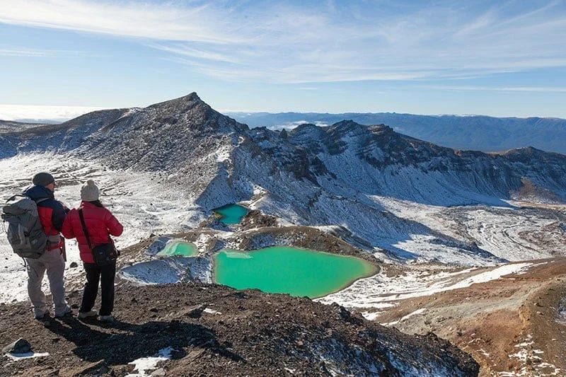 Two hikers overlooking volcanic craters with turquoise lakes in a snow-covered mountainous landscape.