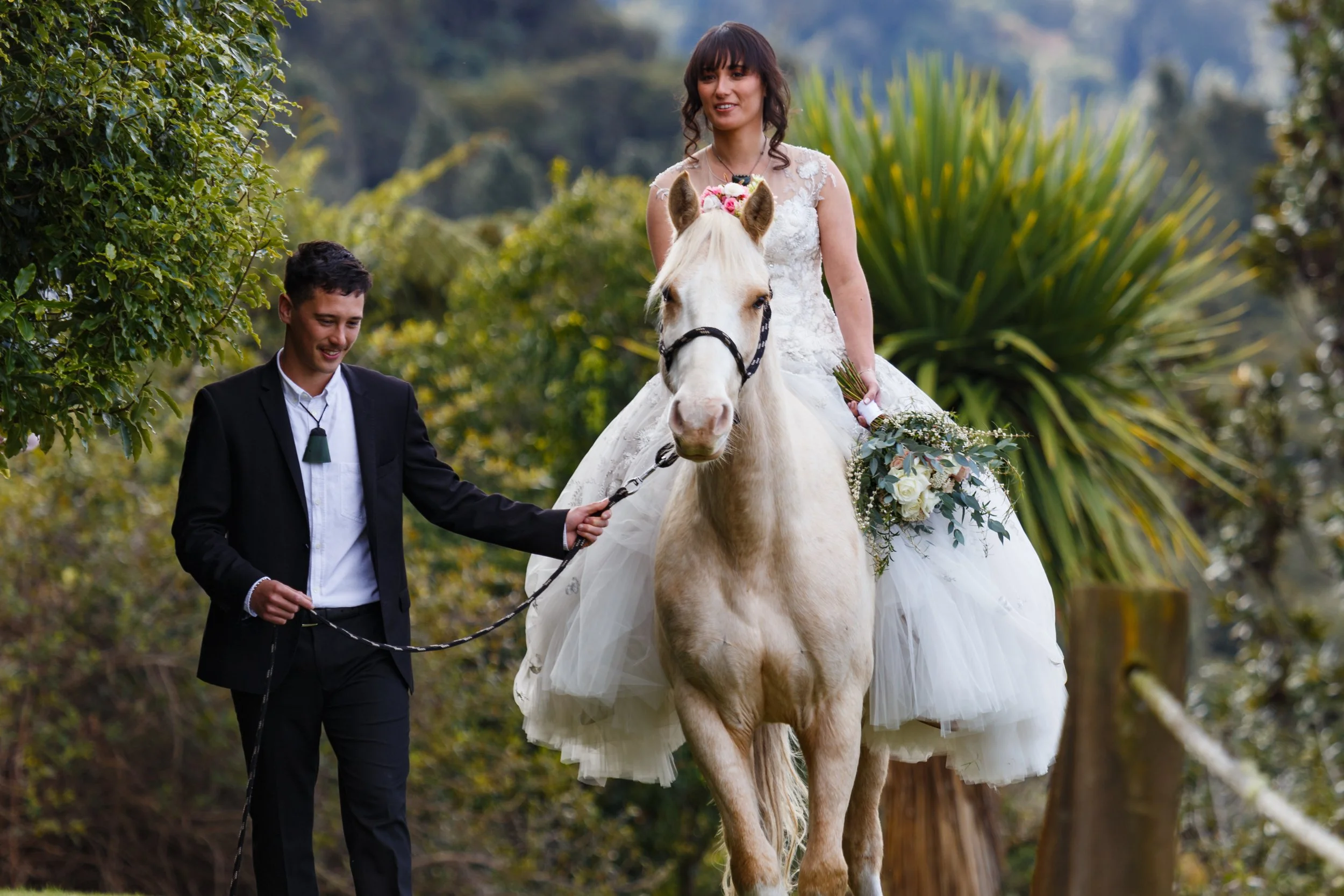 A bride in a white wedding dress riding a white horse, holding a bouquet, while her borther in a black suit walks alongside, helping to guide the horse in an outdoor setting to the groom.