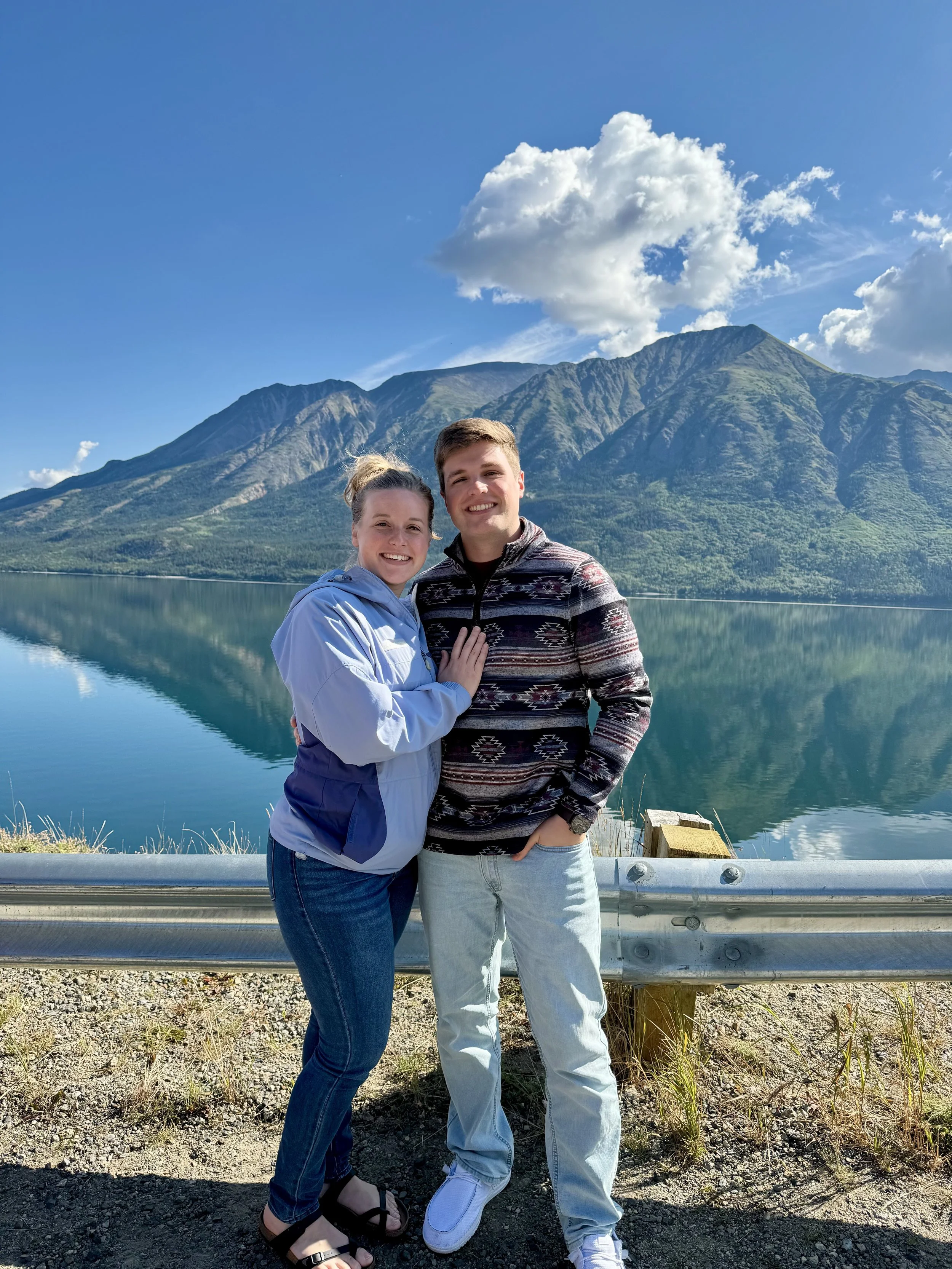 A smiling couple standing together outdoors in front of a mountain range reflecting on a lake, with a guardrail and grassy area in the foreground and partly cloudy sky above.