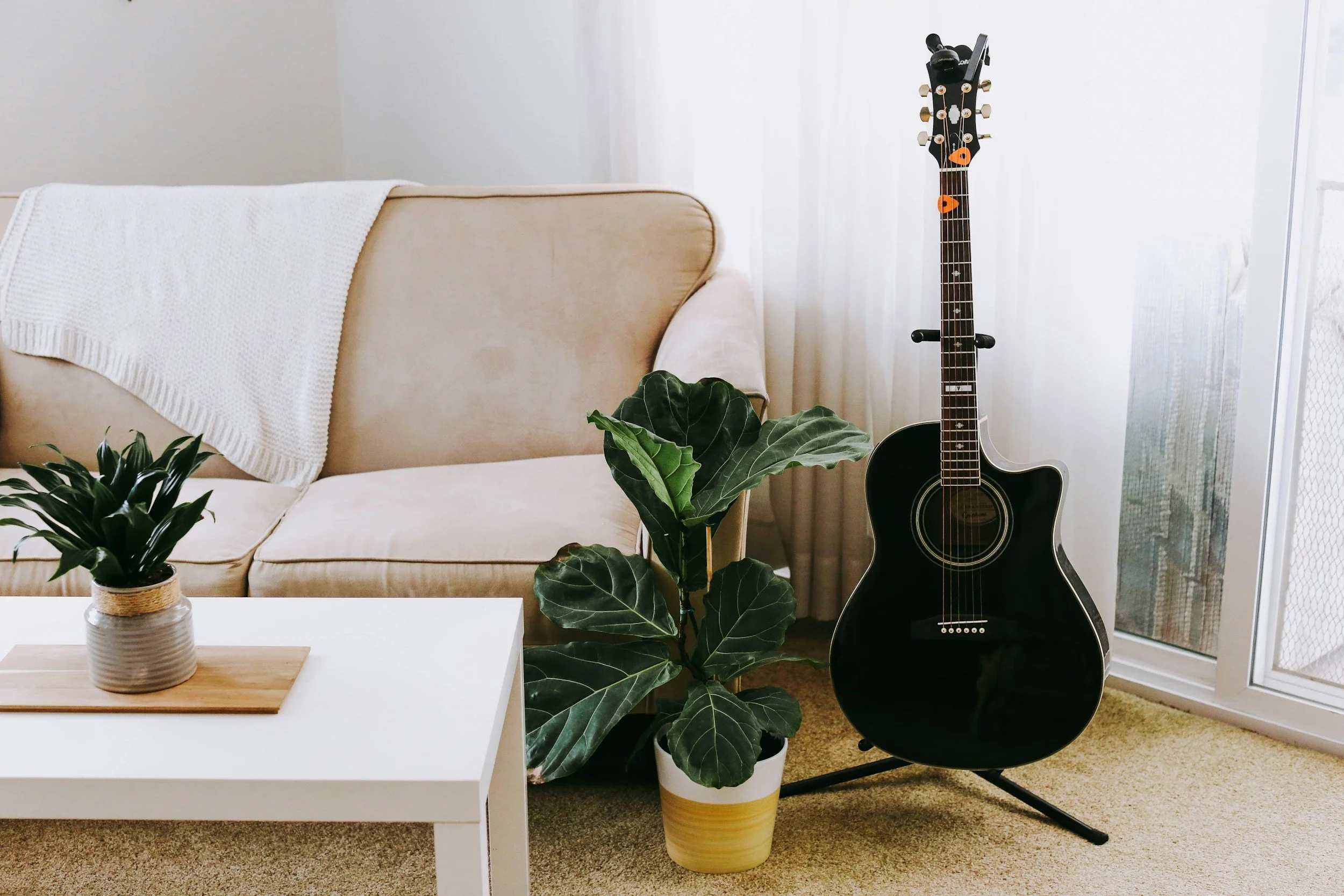 Living room with a beige couch draped with a white blanket, a white coffee table with a potted plant, a black acoustic guitar on a stand, and several larger potted plants, with a window covered by a sheer curtain.