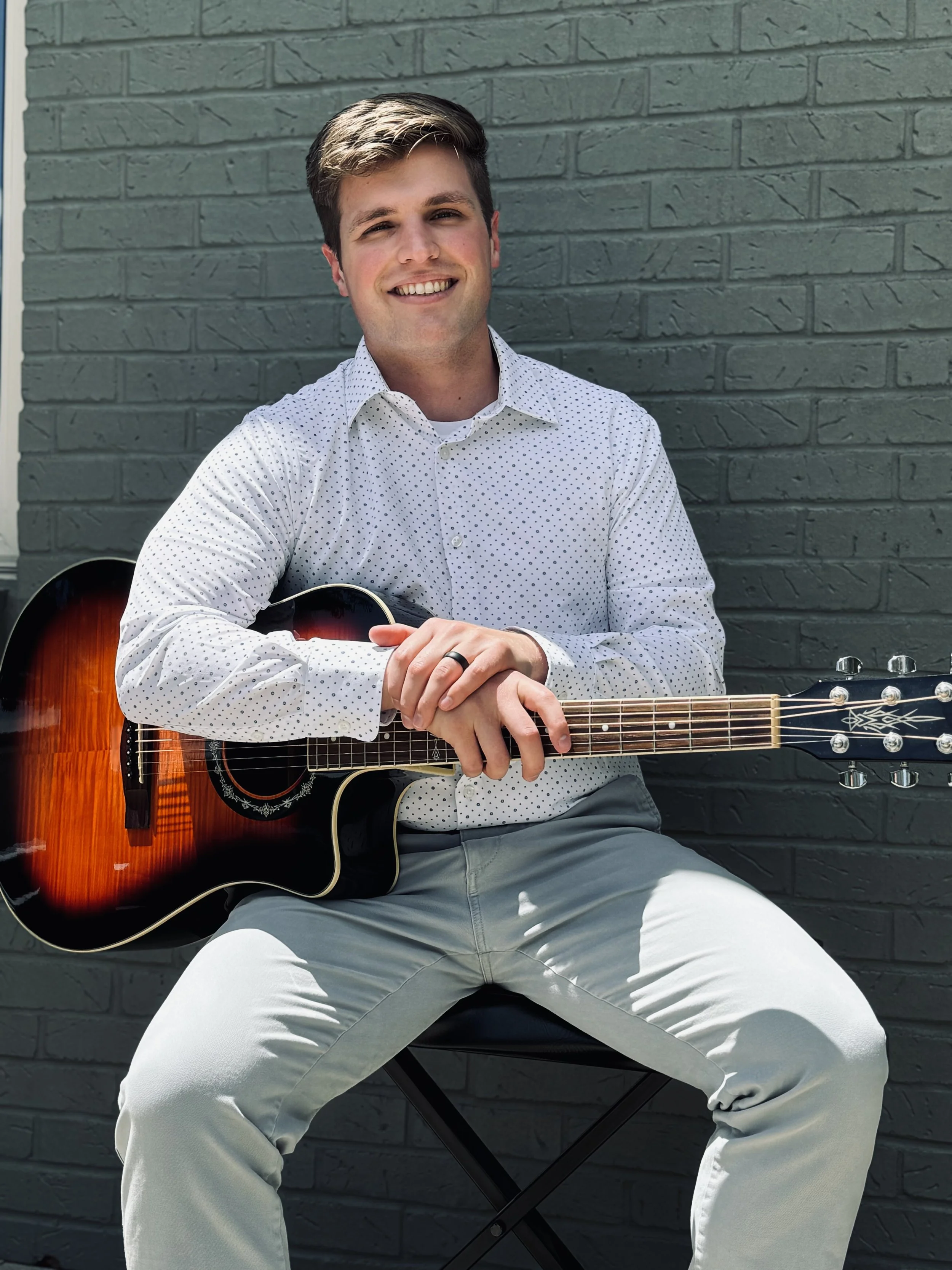 Young man sitting outdoors against a gray brick wall, smiling, holding an acoustic guitar, wearing a white polka dot shirt and light gray pants.