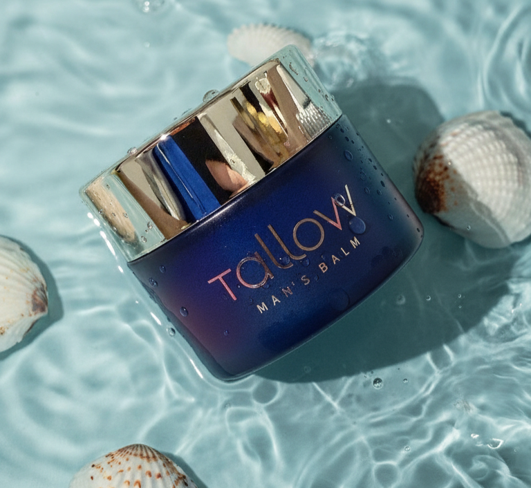 A jar of TalloVY man's balm floating in water surrounded by seashells.