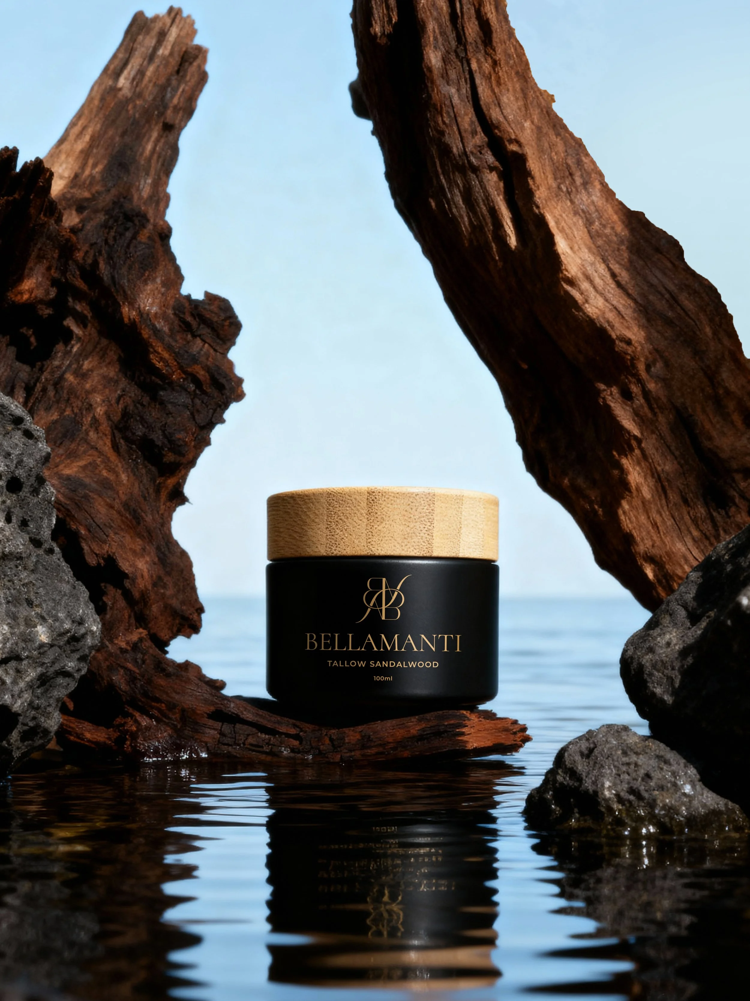 A black jar of Bellamanti Tallow Sandalwood cream with a wooden lid, placed on a piece of driftwood surrounded by rocks and water, with a light blue sky in the background.