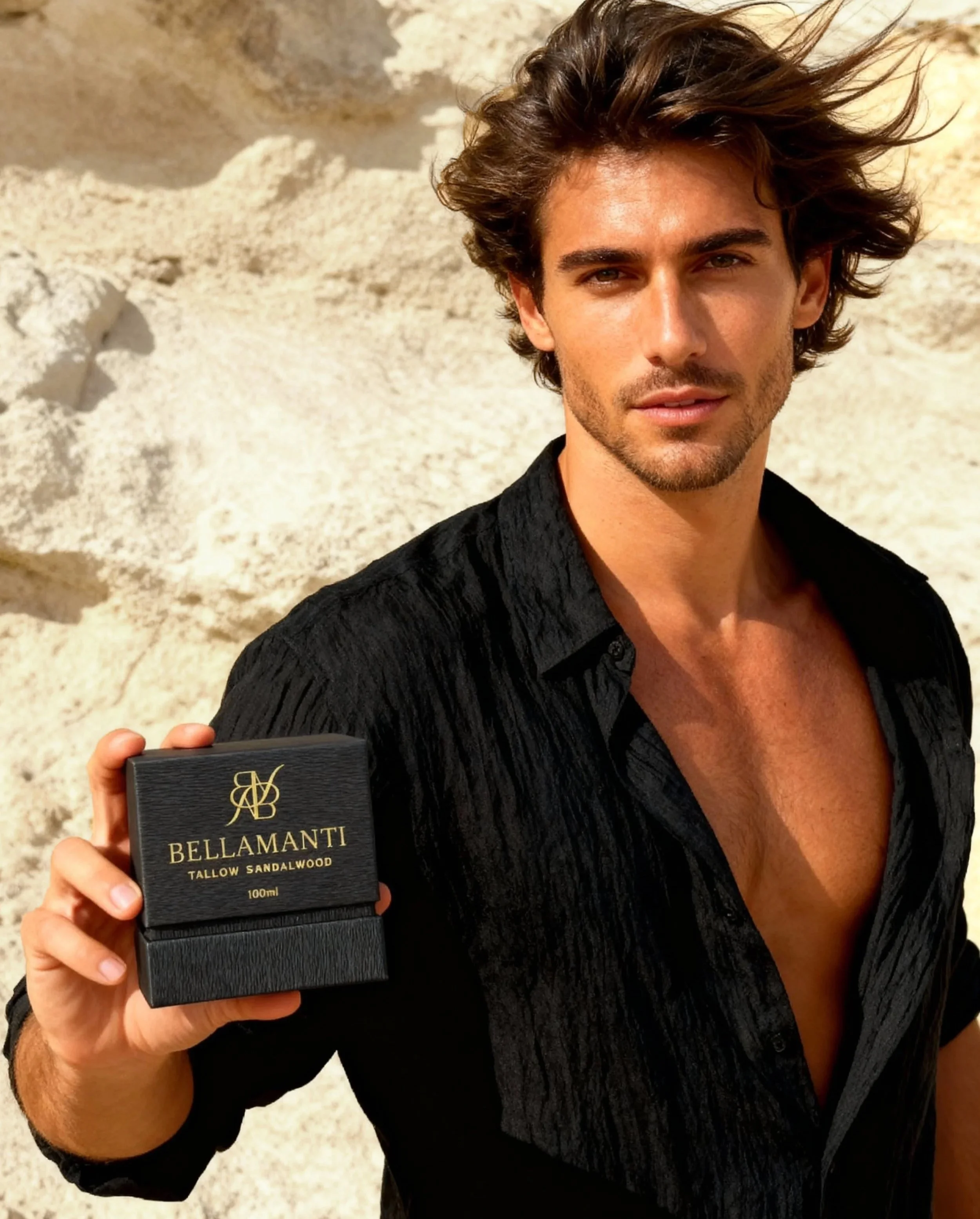 A young man with brown, wavy hair and a light beard, wearing a partially unbuttoned black shirt, standing outdoors against a beige rocky background, holding a black box with gold lettering that says 'Bellamanti Tallow Sandalwood 100ml'.