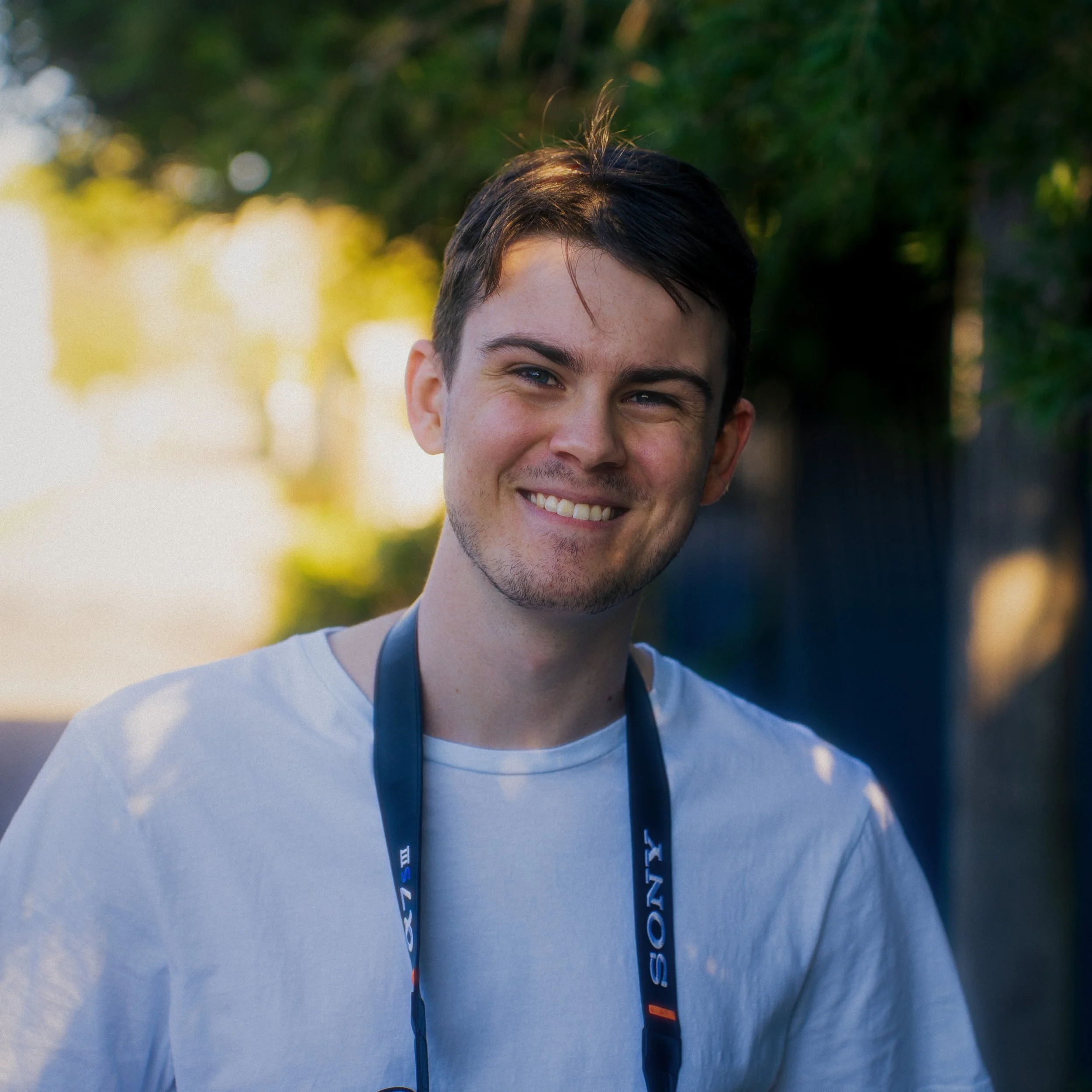 A smiling young man with dark hair wearing a white t-shirt and a Sony camera strap around his neck outdoors with sunlight and trees in the background.