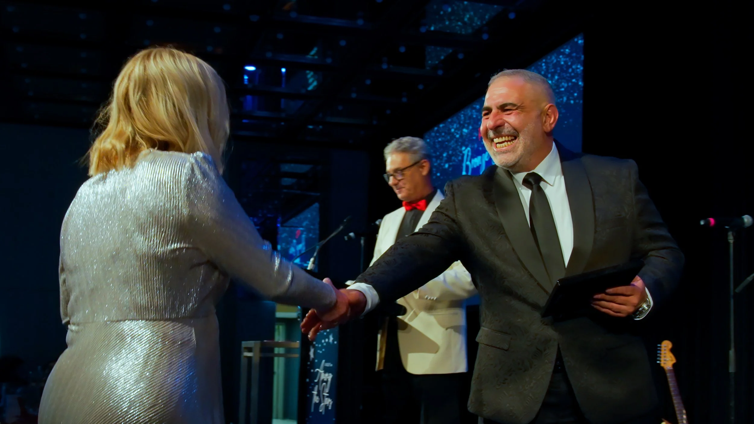 A man with glasses and a beard, wearing a black polo shirt with logos, sitting in a room with a window and unfinished wall.