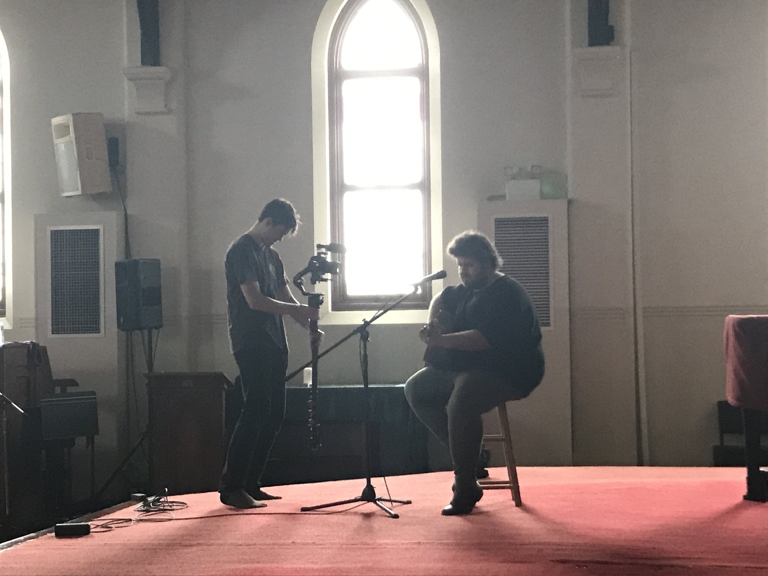 A person is sitting on a chair and holding a guitar or ukulele while being filmed by a cameraman in a room with a high window and soft lighting.