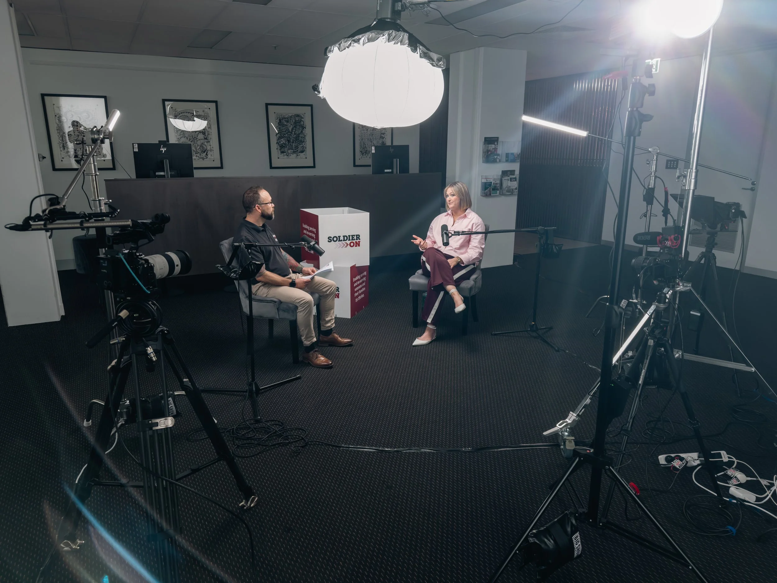 An interview setup with two people, a man and a woman, seated across from each other in a studio. There is a large overhead light, professional camera equipment, microphones, and a box with the words "Soldier ON" behind the man.