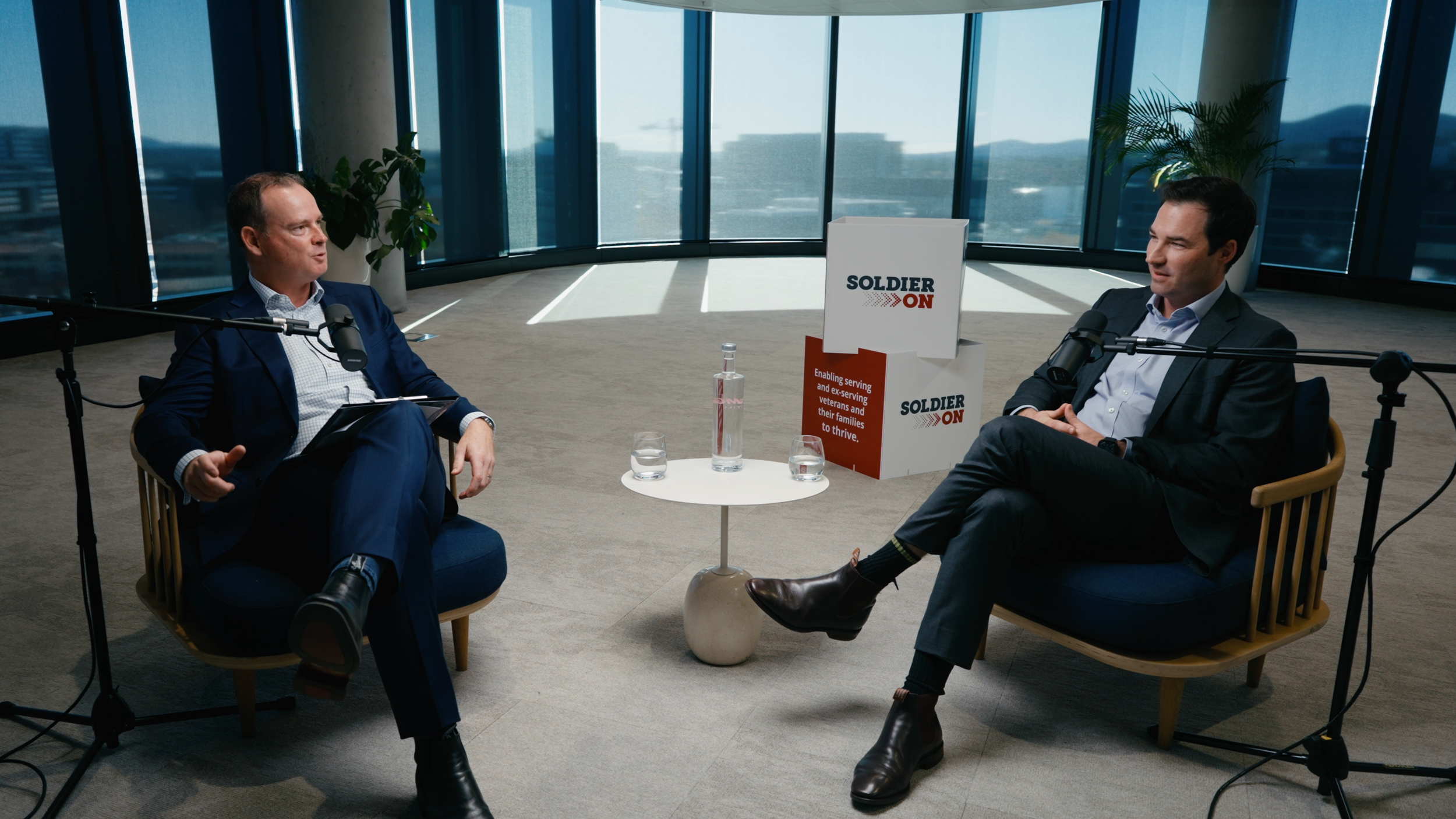Two men in suits having a recorded interview or conversation in a modern office space with large windows, a sign that says 'SOLDIER ON', and a small table with water bottles between them.