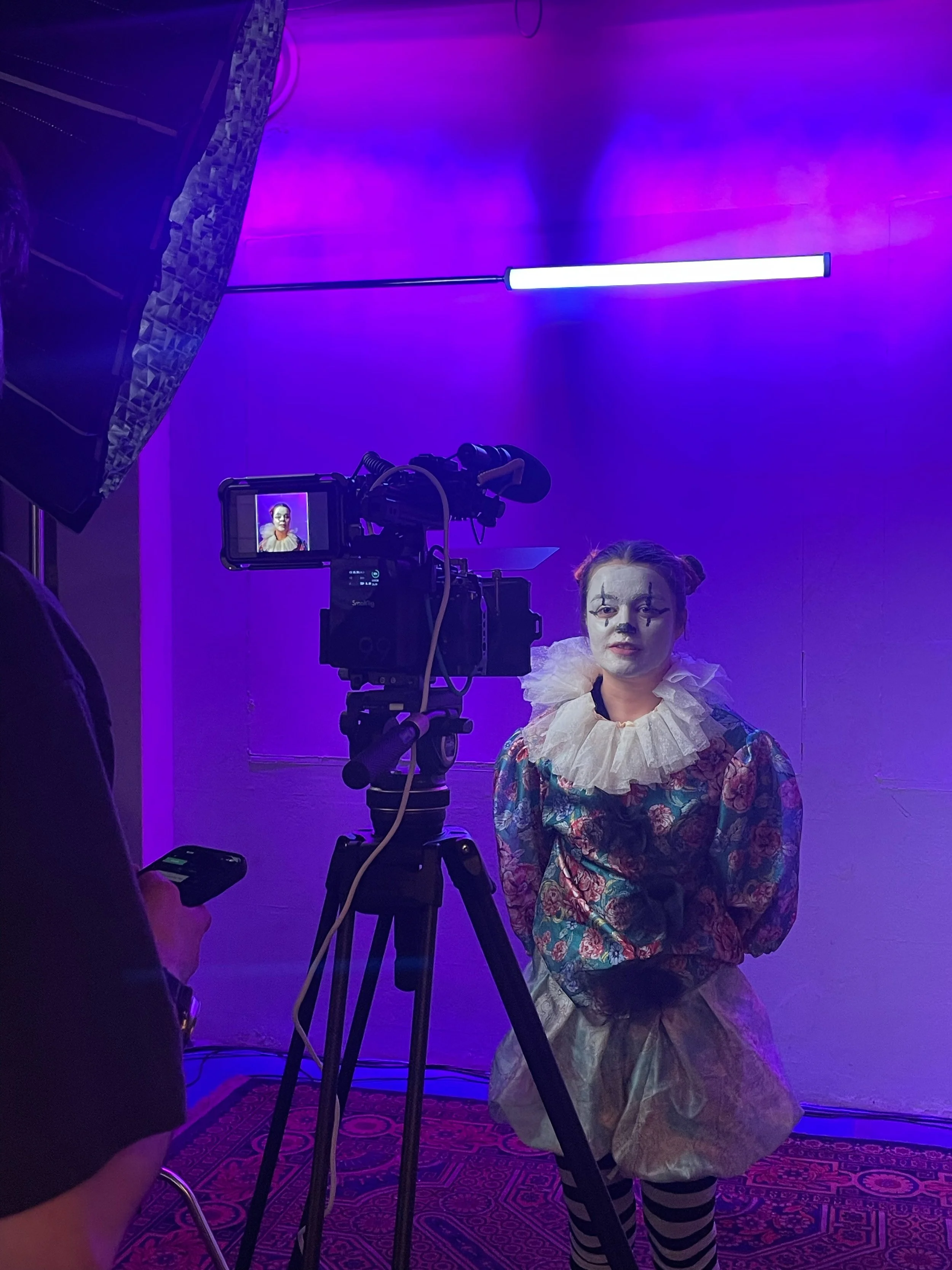 Clown wearing colorful, floral-patterned costume with ruffled collar, makeup with white face paint, painted eyebrows and lips, and striped knee-high socks, standing in front of a purple-lit background while being filmed by a camera operator.