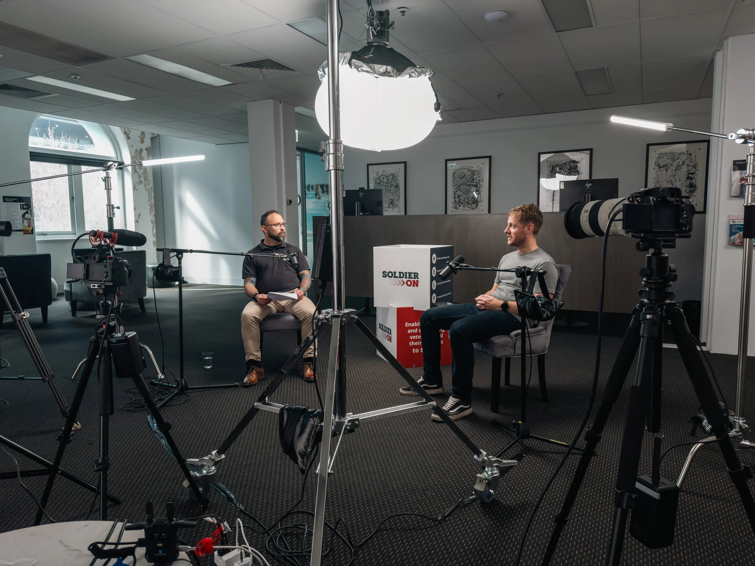 Two men participating in an interview or podcast recorded in a studio with cameras and lighting equipment, with framed artwork on the wall in the background