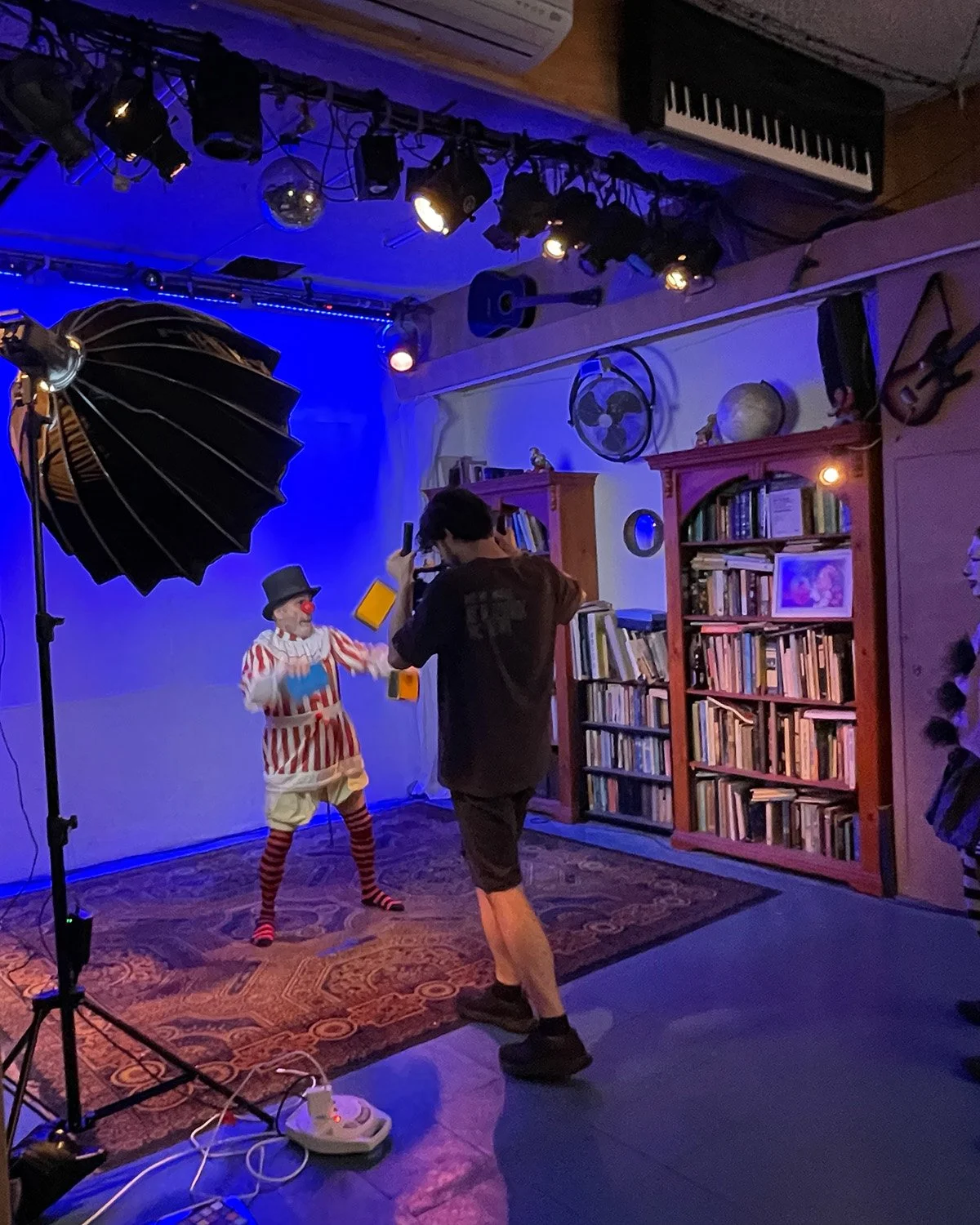 A clown with a gray hat, red nose, and colorful striped shirt performs for a man taking photos in a cozy room with bookshelves, a fan, and a guitar on the wall, under stage lighting.