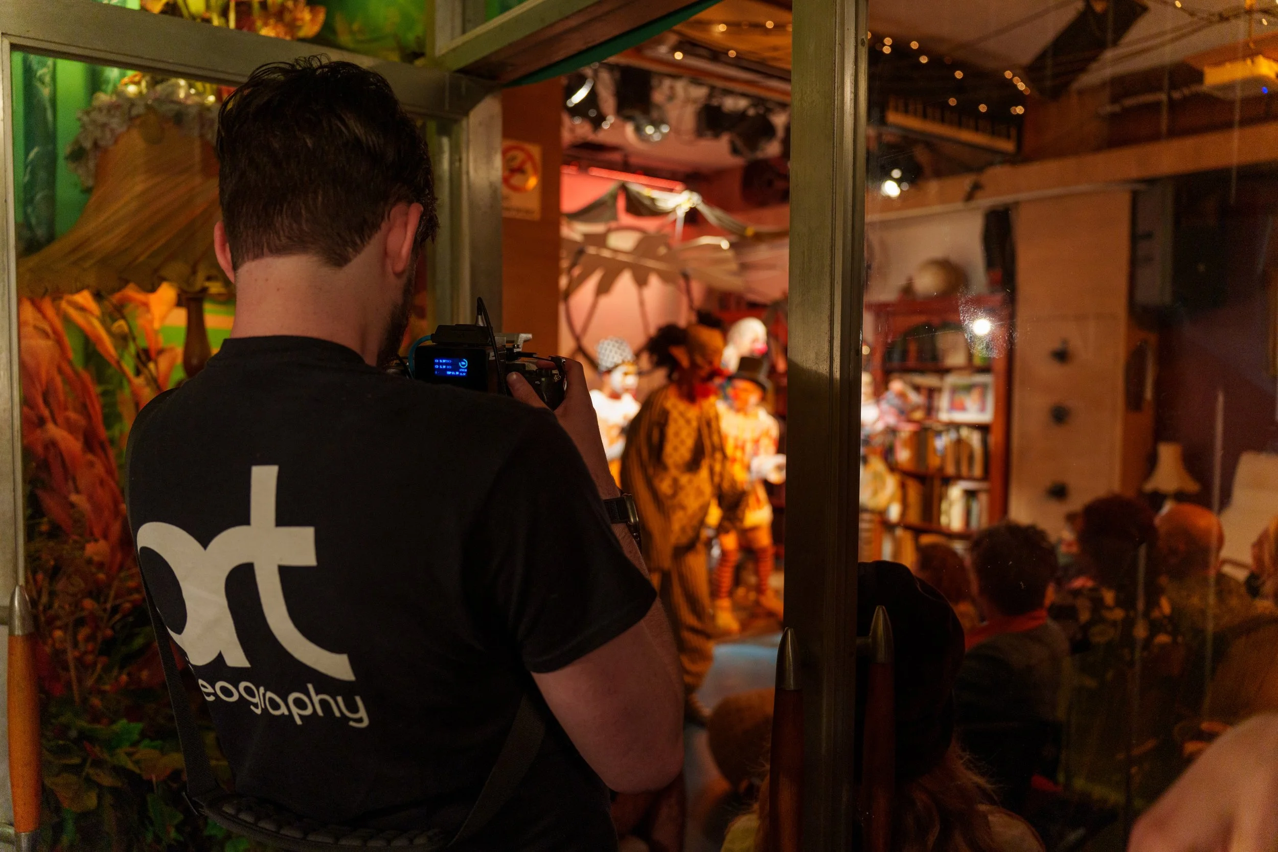 A person with short dark hair taking a photograph of a stage scene through a doorway at an indoor event. The scene includes people dressed in colorful costumes, resembling a theatrical or performance setting.