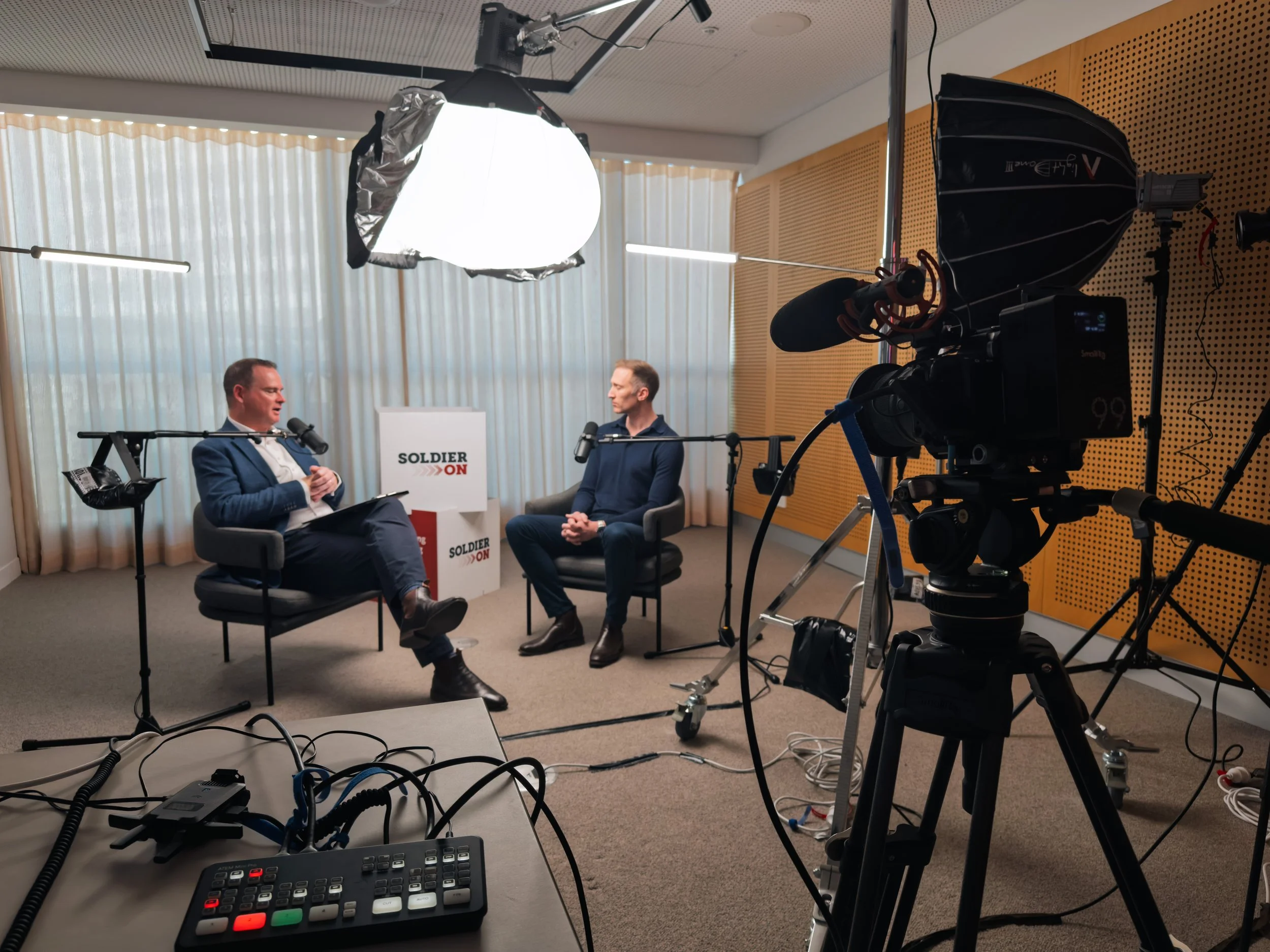 Two men in suits having a conversation in a modern office with large windows, sitting on armchairs with a small table between them, microphones set up for recording.