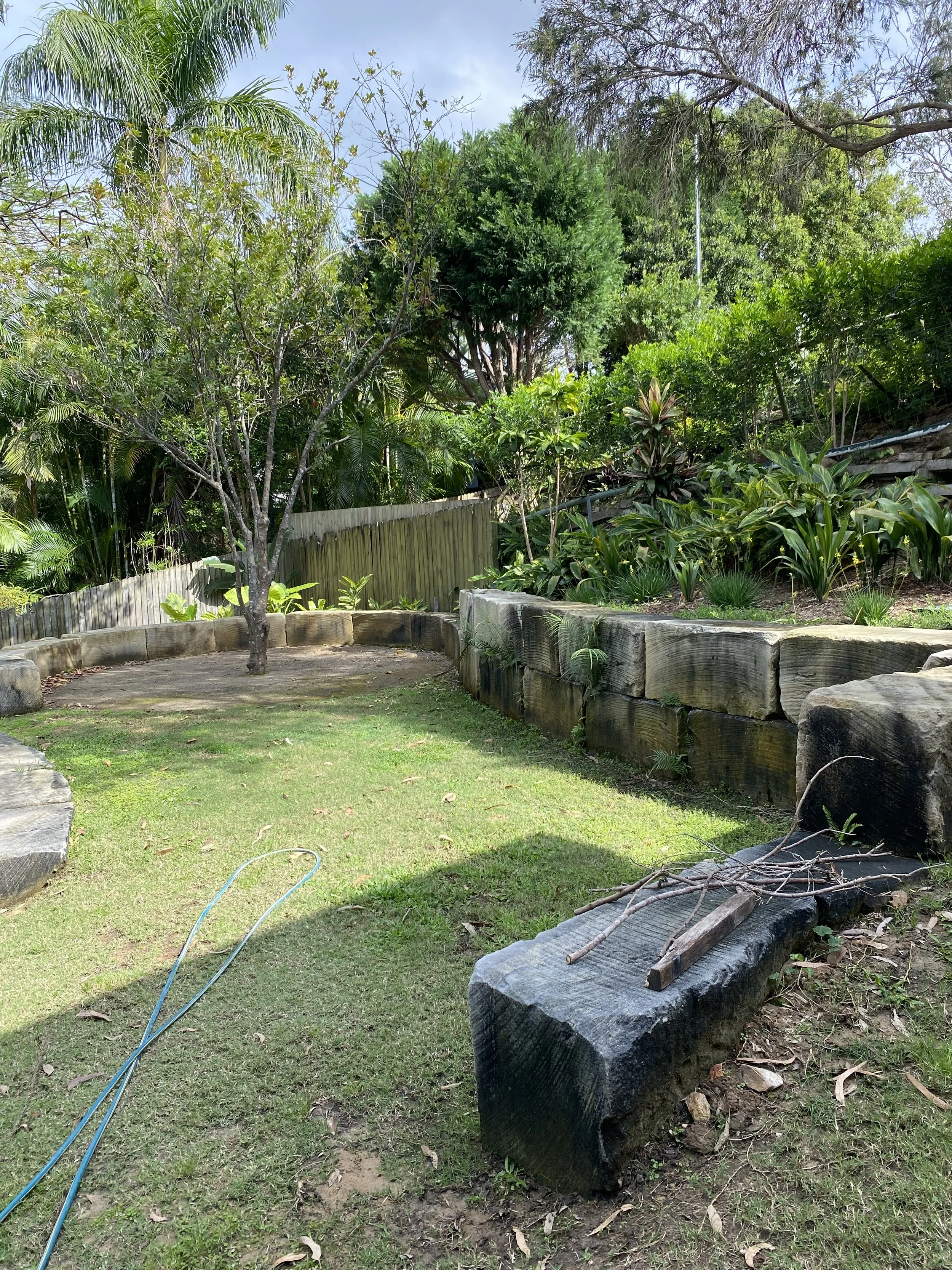 A backyard garden with a grassy area, trees, and plants, enclosed by a wooden fence. There are large rocks and logs along the edges, with some garden tools or sticks on the ground.