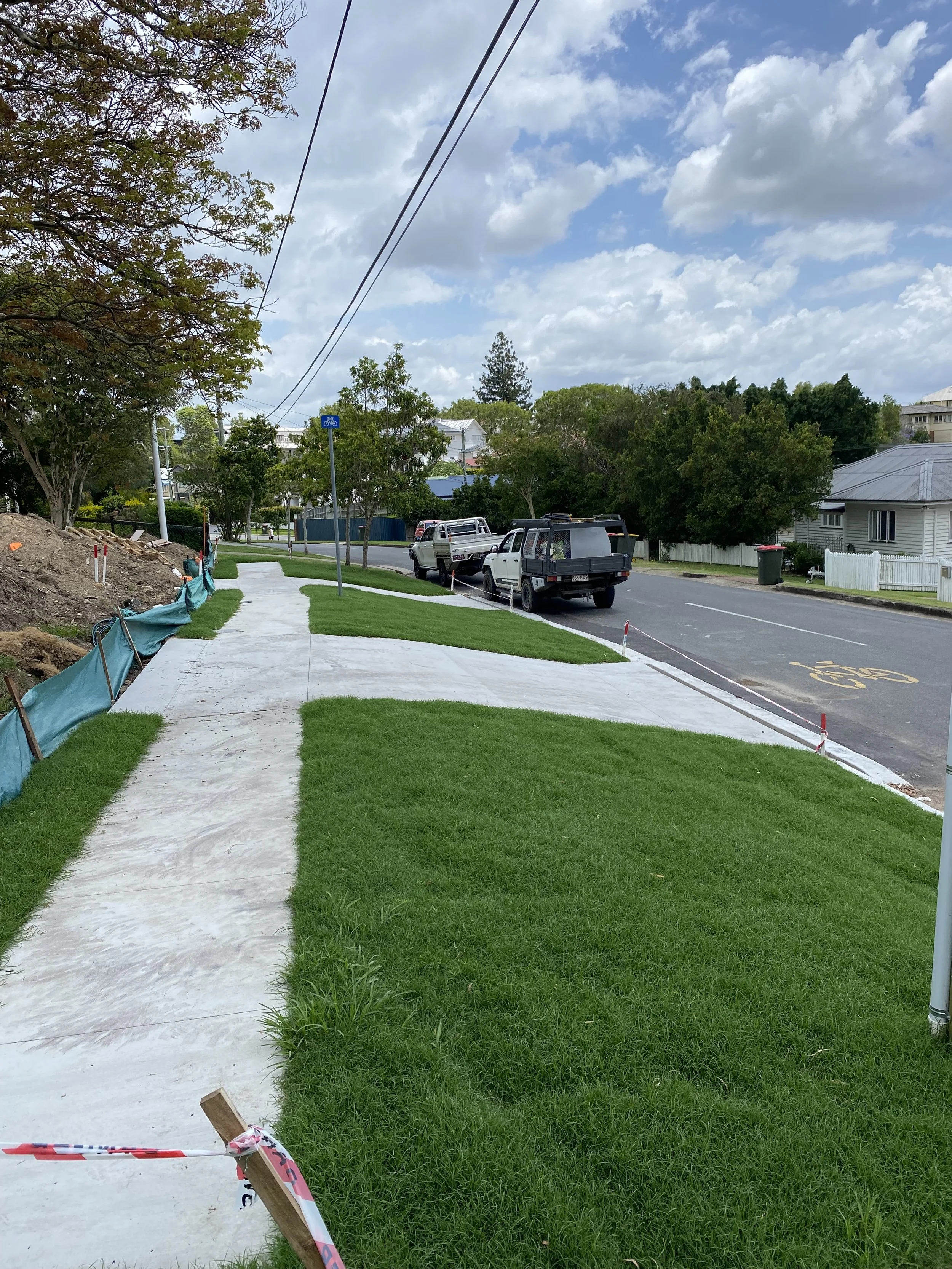 Sidewalk under construction with a grassy strip on both sides, parked trucks on the street, trees lining the sidewalk, and a partly cloudy sky.