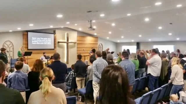 Church congregation gathered, some standing and some seated, with a prominent cross on the wall and a worship band leading in front of screens displaying lyrics.