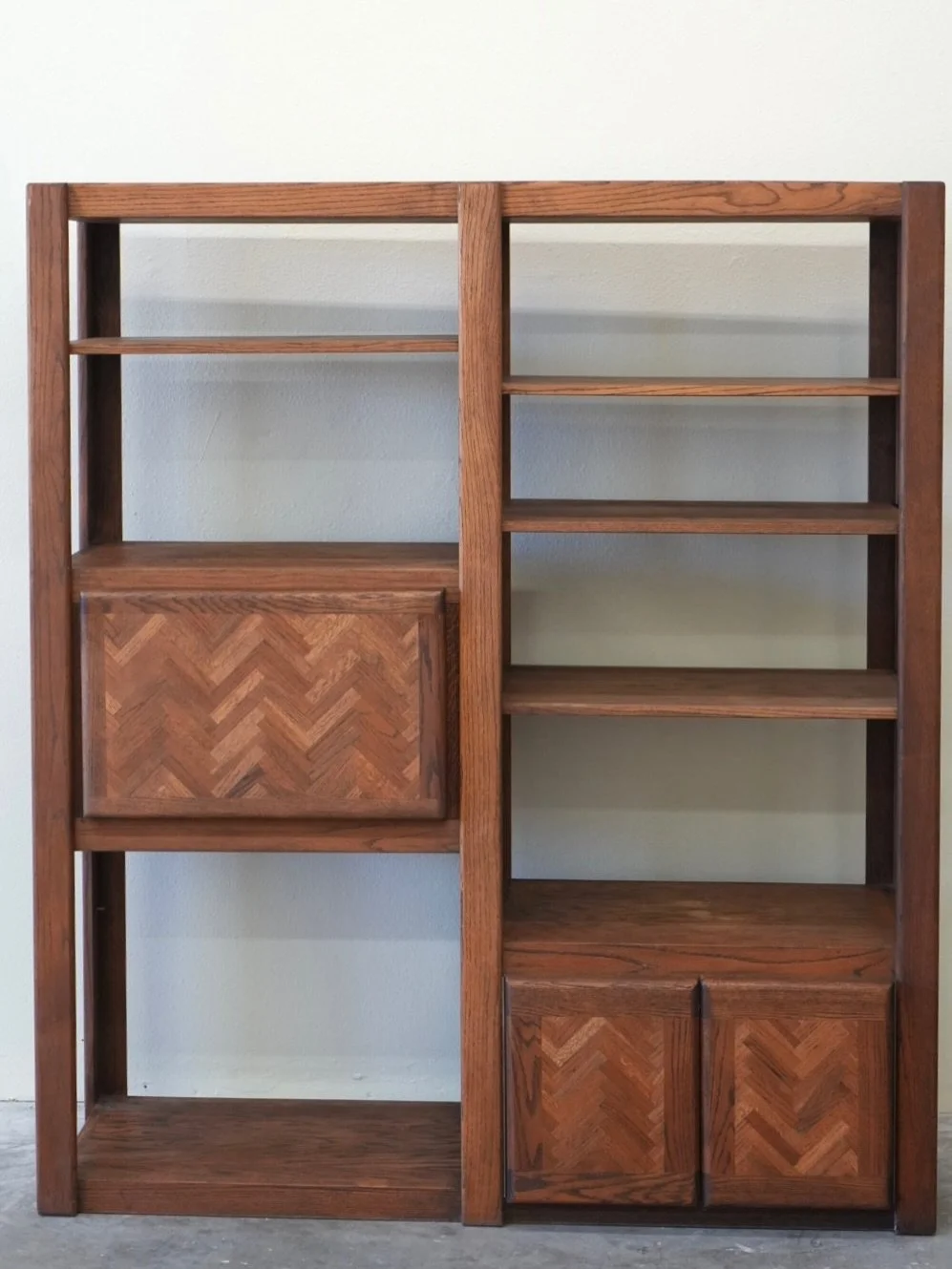 A solid oak wall unit, designed in the style of Lou Hodges. The bookcase features a timeless design with open shelving, a distinctive herringbone-patterned cabinet door, and additional storage with double doors below. 

65&rdquo;x19.5&rdquo;x76&rdquo