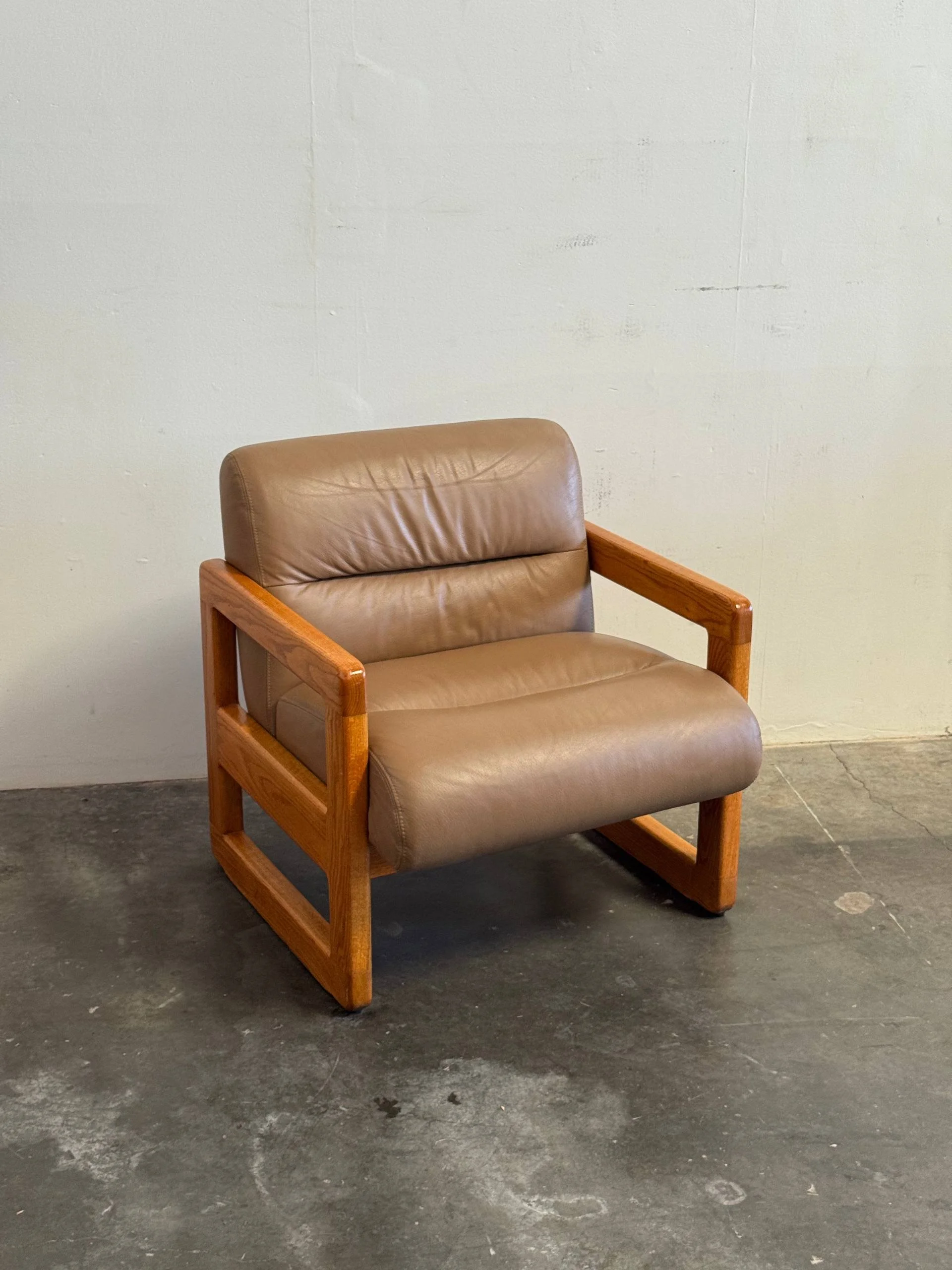 A brown leather armchair with a wooden frame placed against a plain off-white wall, on a concrete floor.