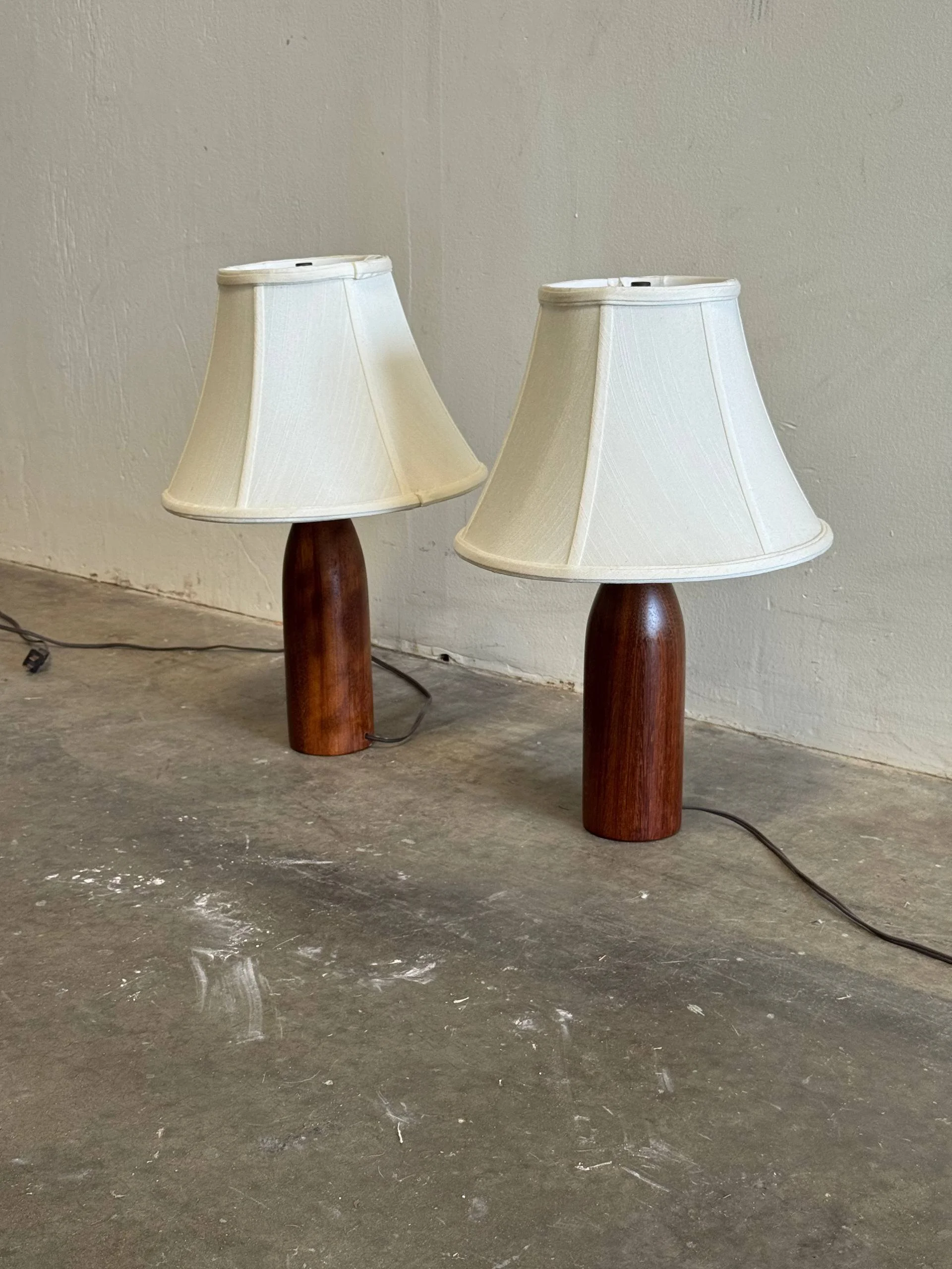 Two table lamps with white shades and wooden bases on a concrete floor against a plain wall.
