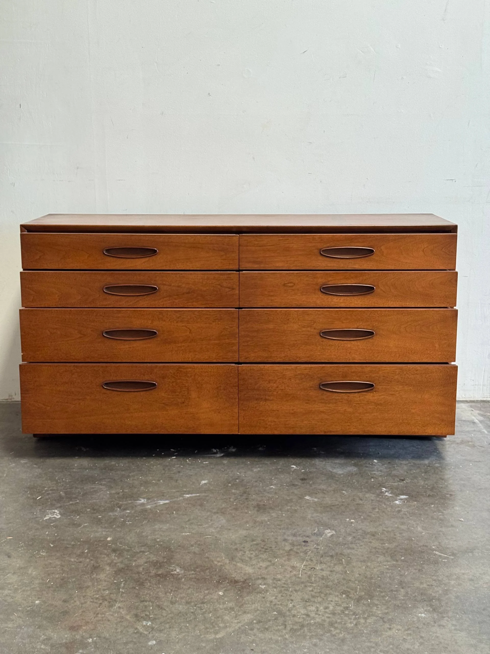 A mid-century modern wooden dresser with six drawers, set against a plain white wall and placed on a concrete floor.