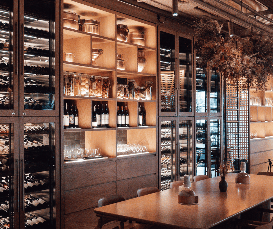 Wine bottles and glasses stored in wooden shelves and wine refrigerators in a modern dining room with a wooden table and decorative vases.