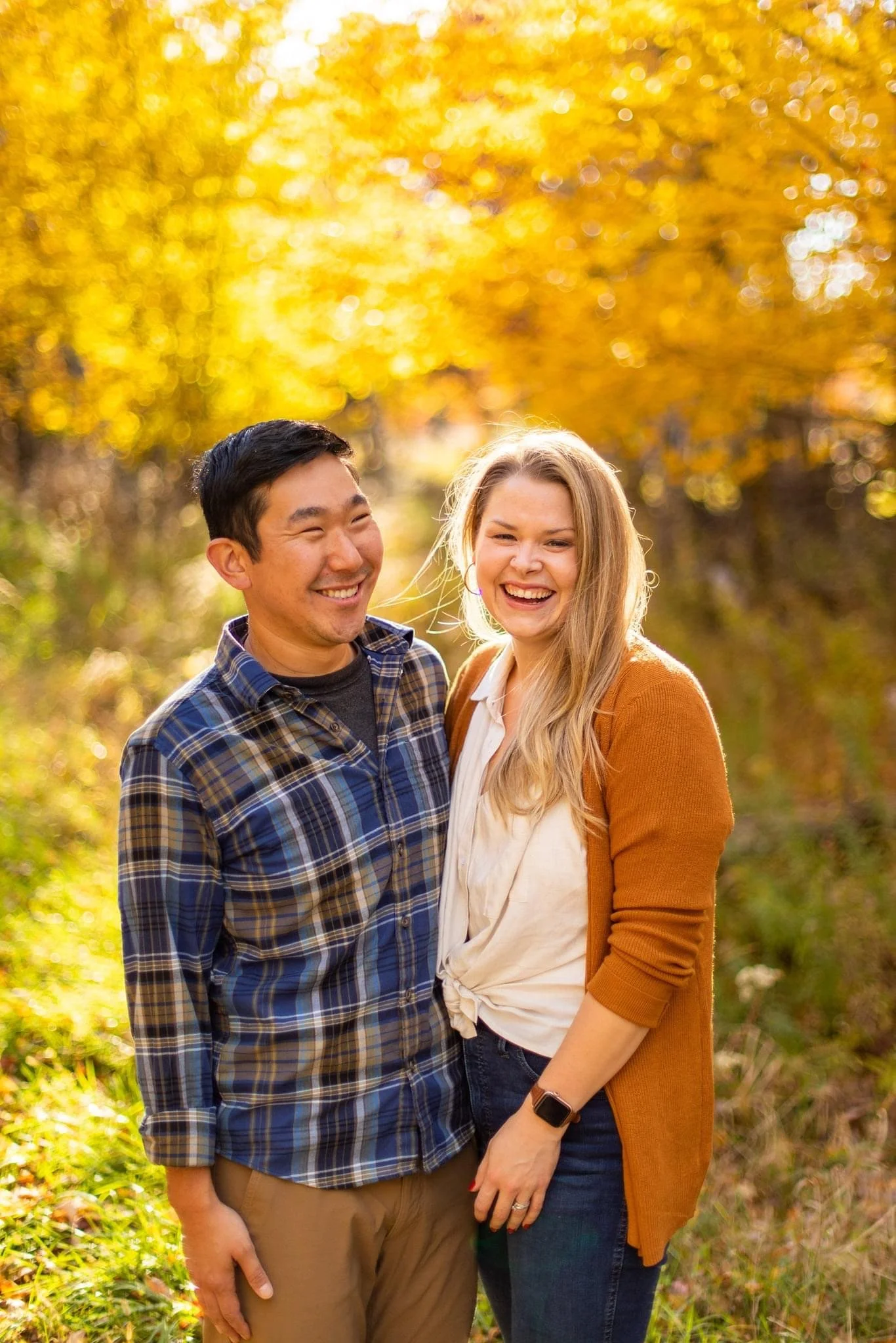 A young man and woman smiling and standing close together outdoors in a park with yellow autumn trees in the background.
