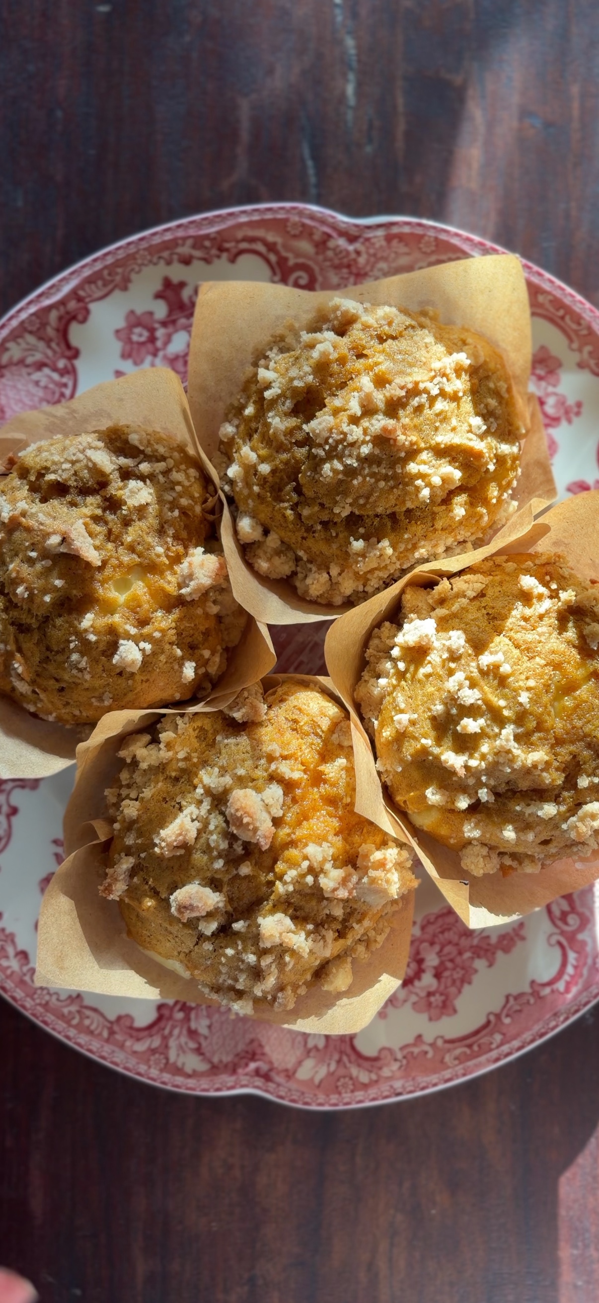 Four crumb-topped muffins in parchment paper liners on a vintage pink and white patterned plate.