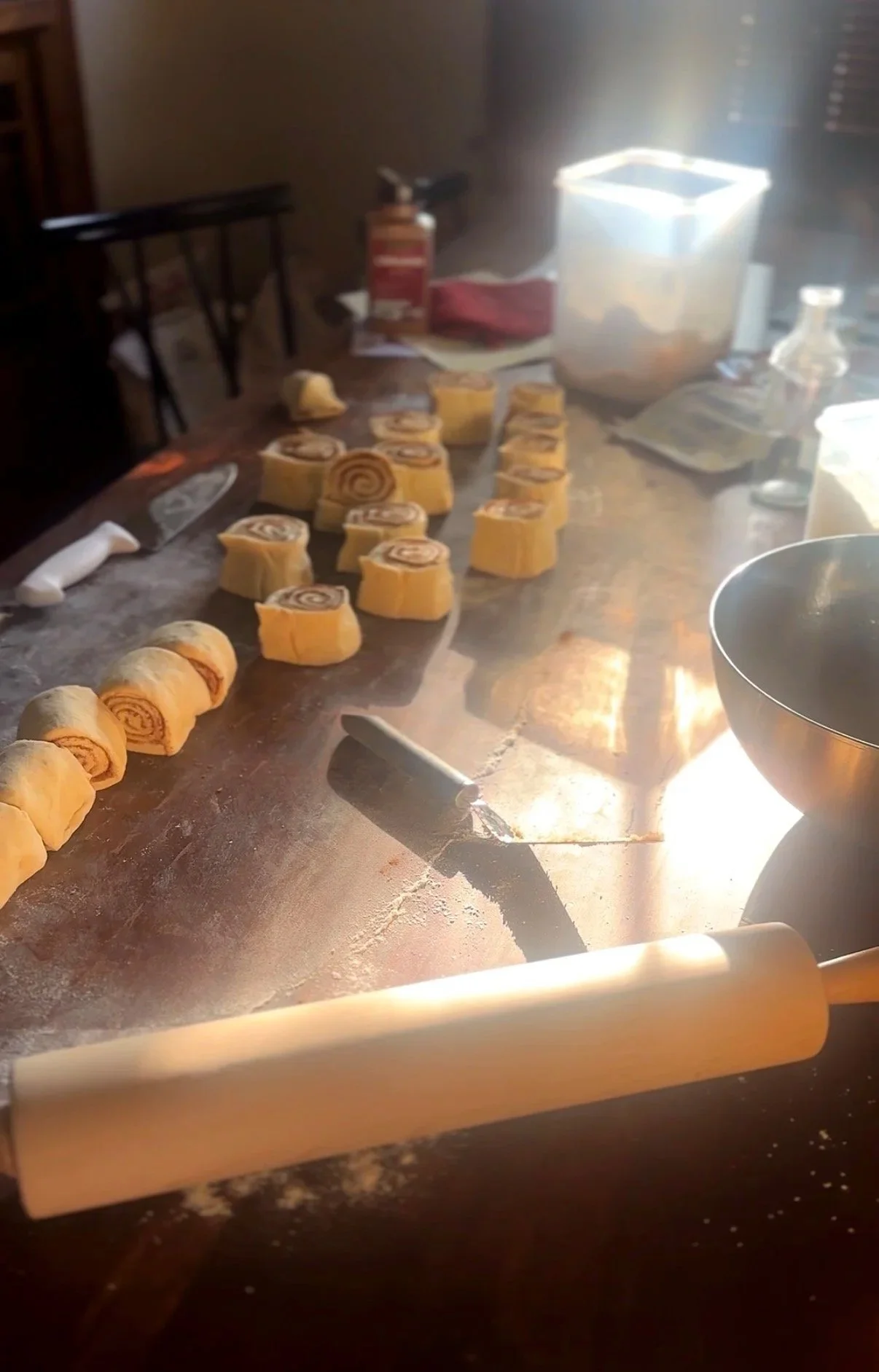 Rolling out dough and preparing cinnamon rolls in a kitchen with a rolling pin, baking tools, and ingredients on the counter, illuminated by bright sunlight.