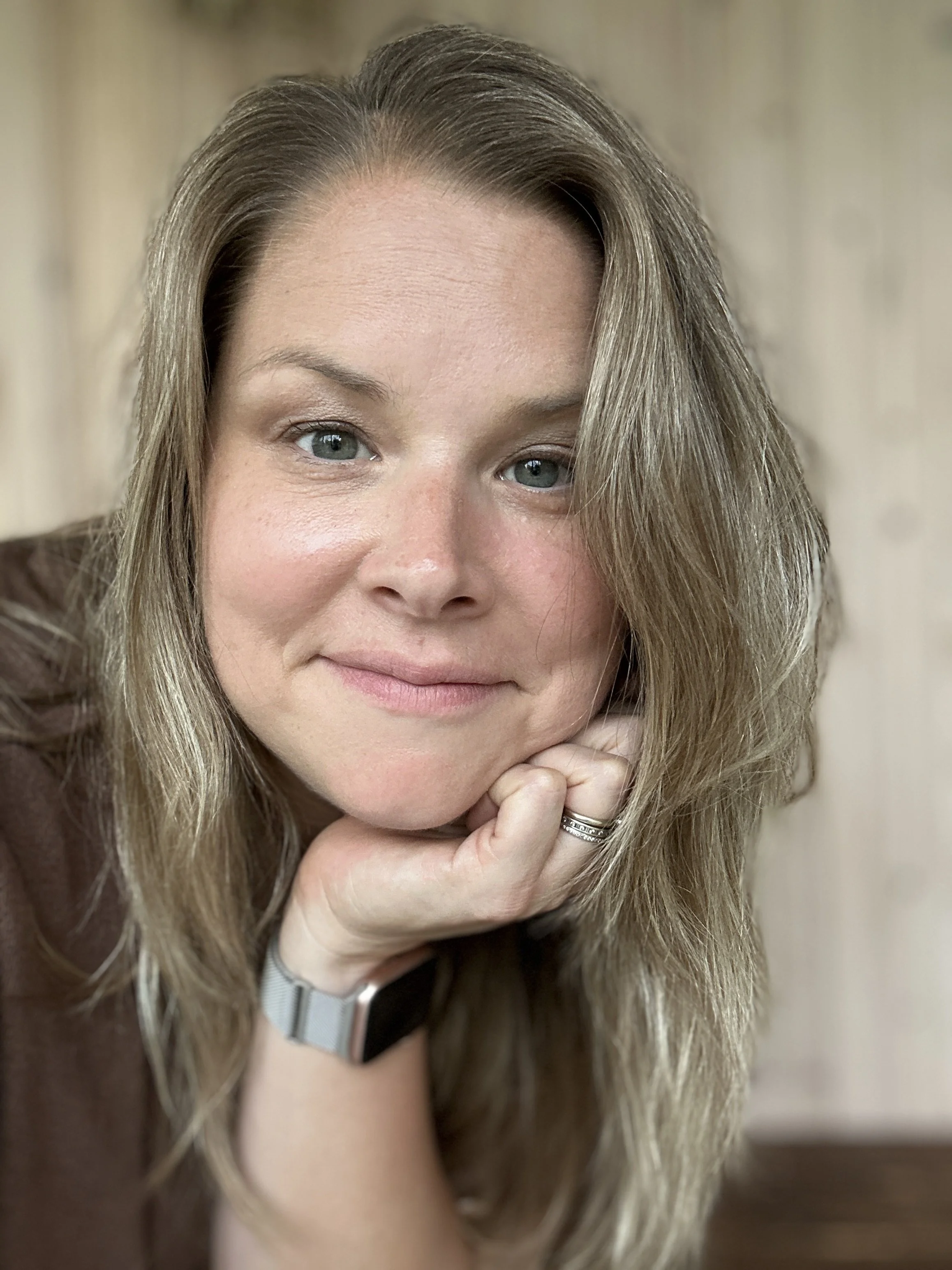 Close-up of a woman with long blonde hair, blue eyes, and light skin, resting her chin on her hand, wearing a ring, and looking at the camera.