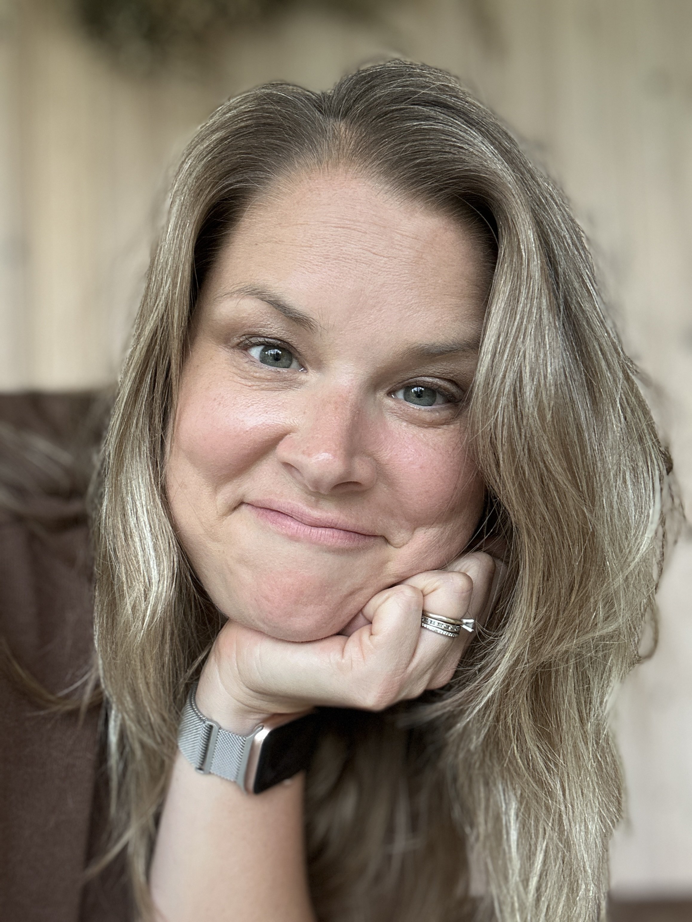 Close-up of a woman with blonde hair, green eyes, and wearing rings, resting her chin on her hand and smiling softly.